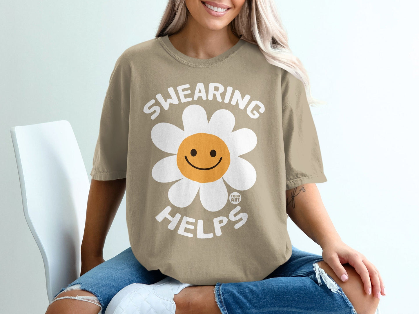 Tan t-shirt with smiling daisy graphic and "SWEARING HELPS" text.