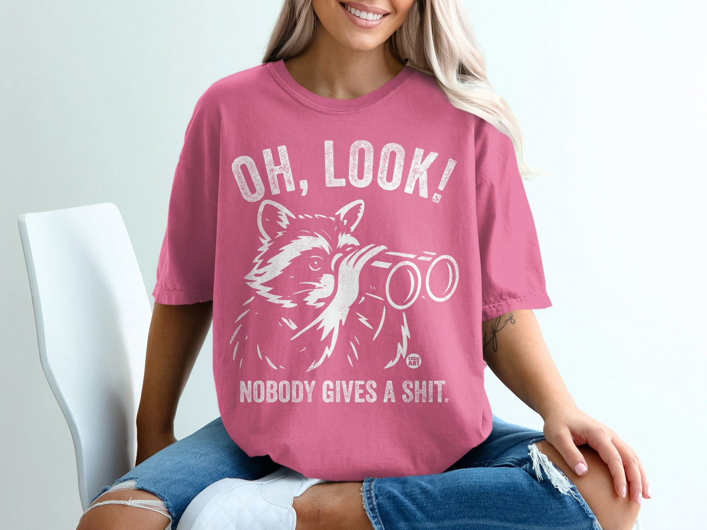 Pink t-shirt with raccoon looking through binoculars and text "OH, LOOK! NOBODY GIVES A SHIT."