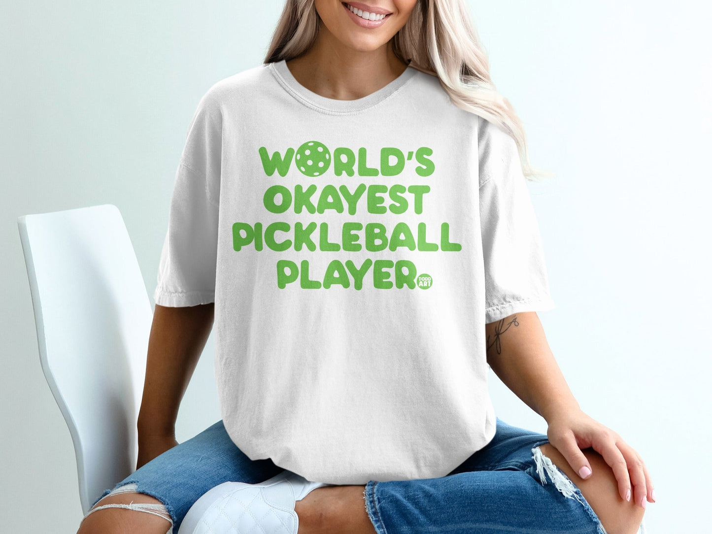 White t-shirt with "WORLD'S OKAYEST PICKLEBALL PLAYER" in green lettering.