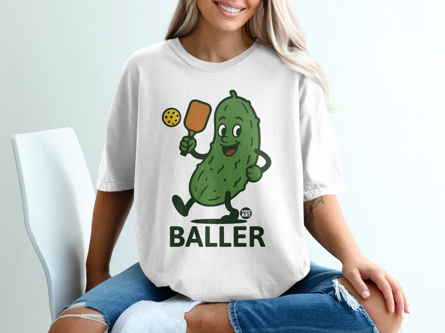 Cartoon pickle playing pickleball on a white t-shirt.