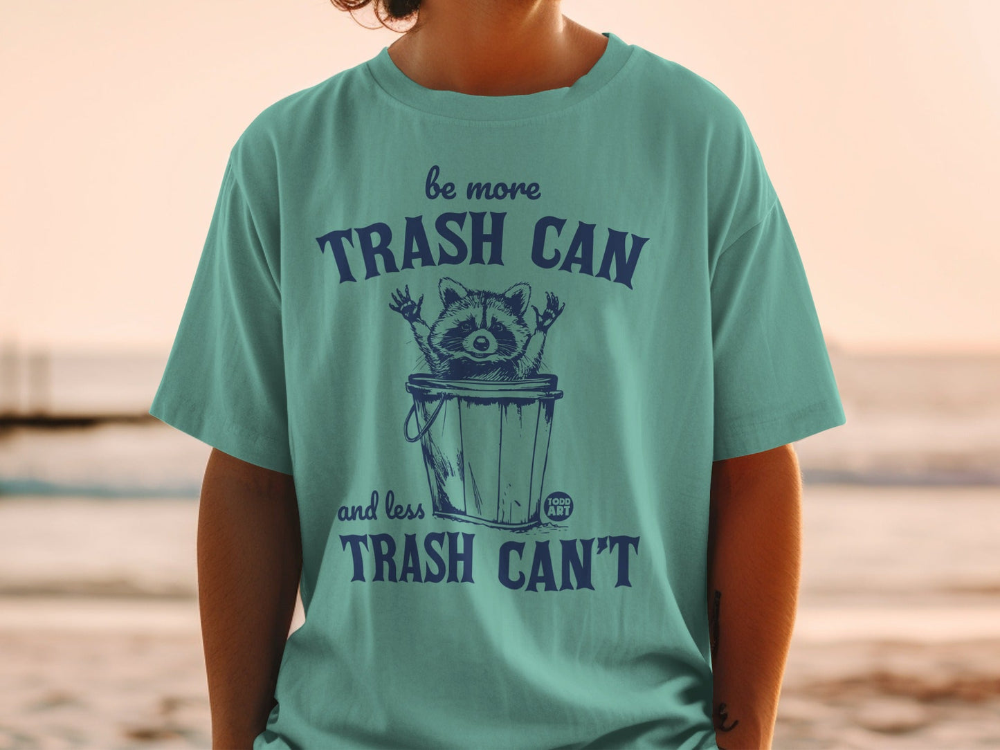 Teal t-shirt with raccoon in trash can and text "Be more trash can and less trash can't".