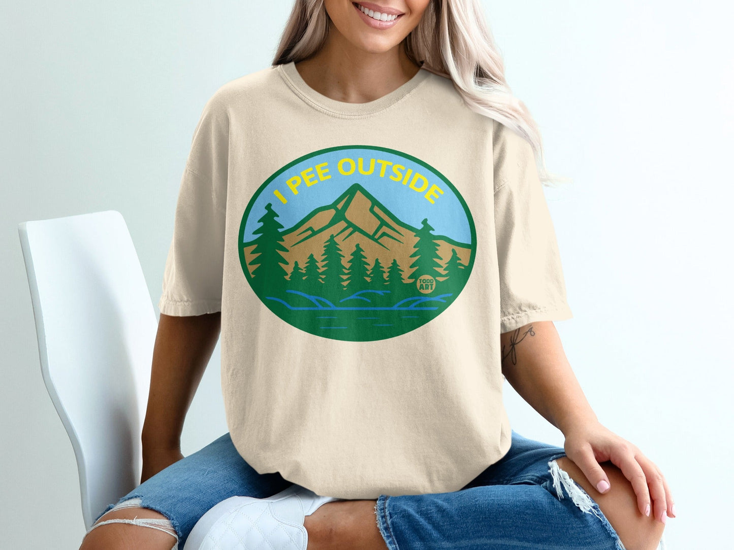 Cream t-shirt with oval graphic of mountains, trees, and lake, text "I PEE OUTSIDE".