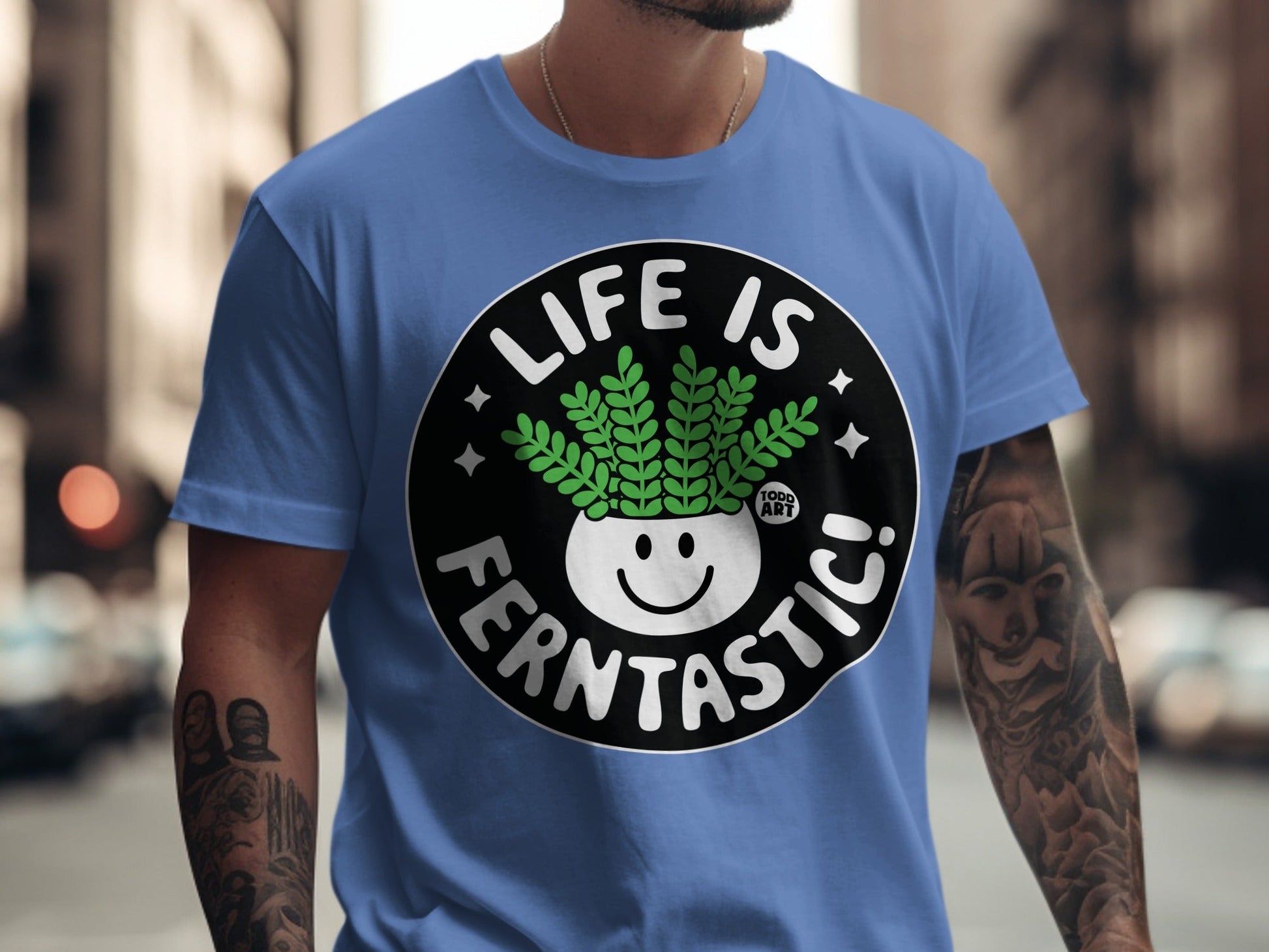 Blue t-shirt with a circular graphic of a smiling plant in a pot that says "LIFE IS FERNTASITC!".