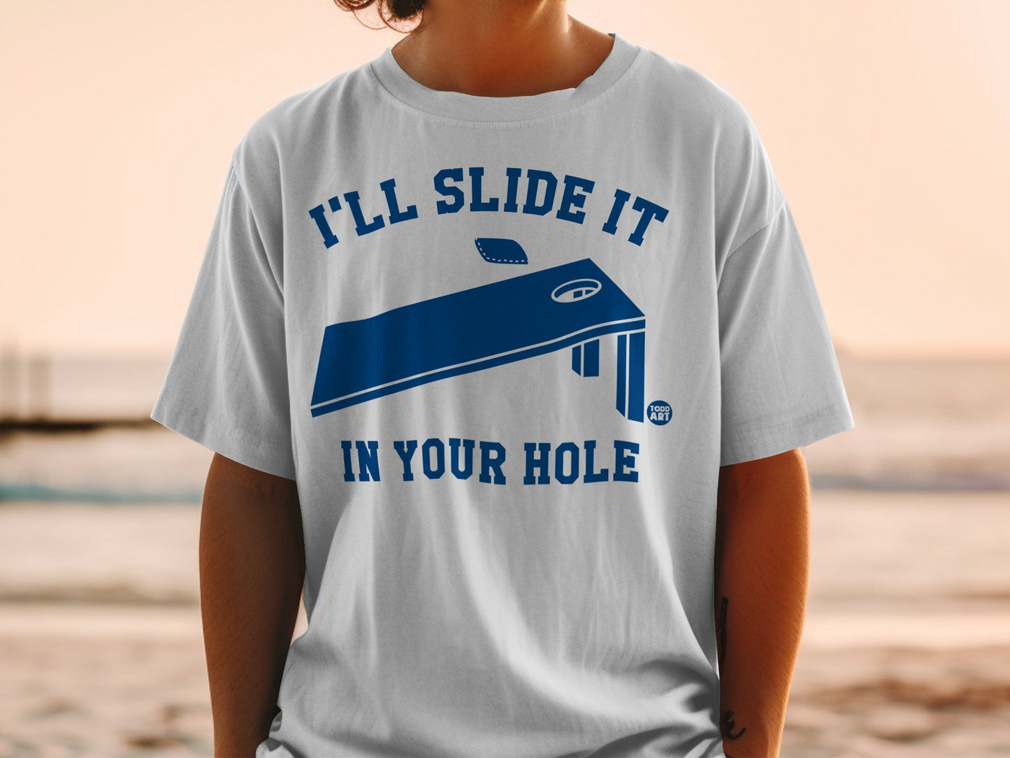White t-shirt with dark blue graphic of a cornhole board and text.