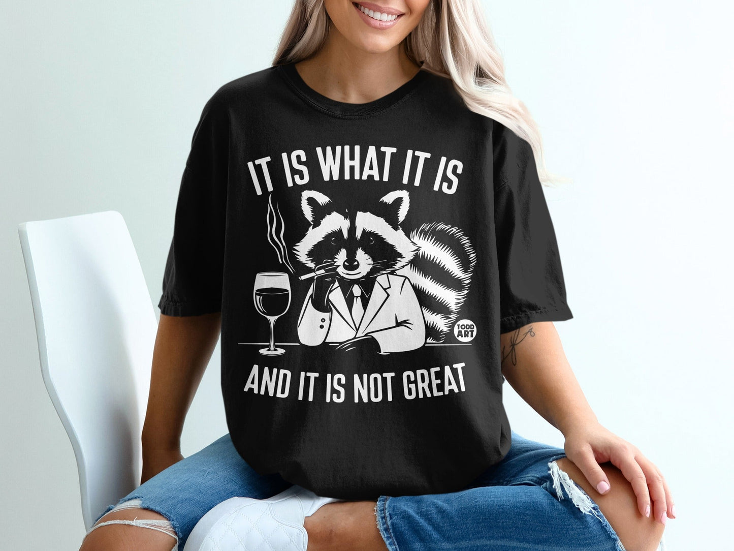 Black t-shirt with raccoon in suit, smoking, holding wine, text IT IS WHAT IT IS AND IT IS NOT GREAT.