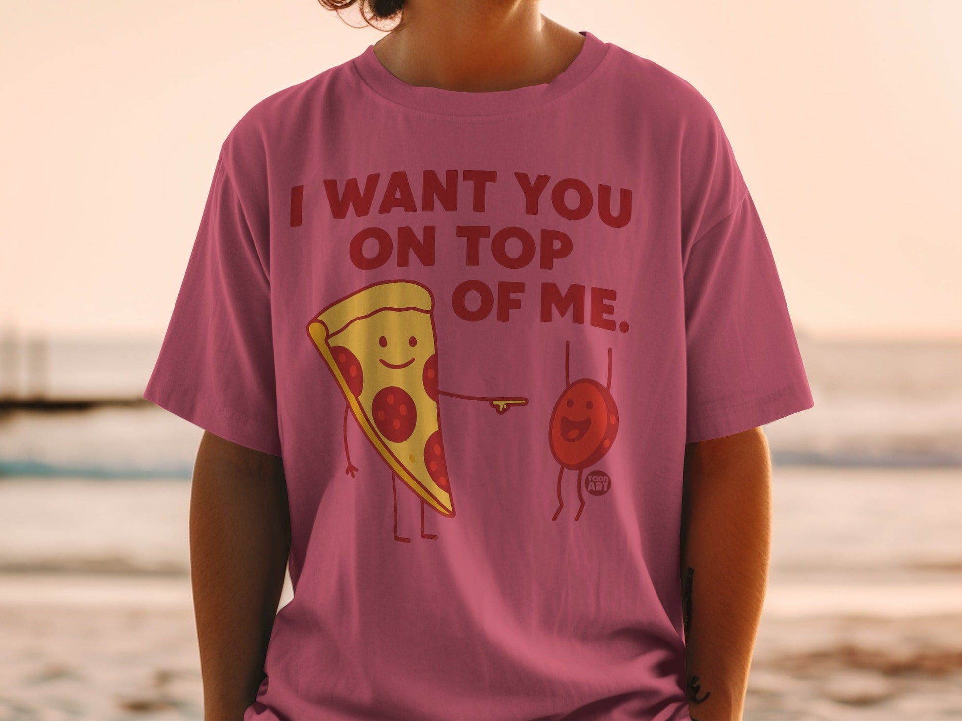 Pink t-shirt with a slice of pizza and a tiny red character saying "I WANT YOU ON TOP OF ME."