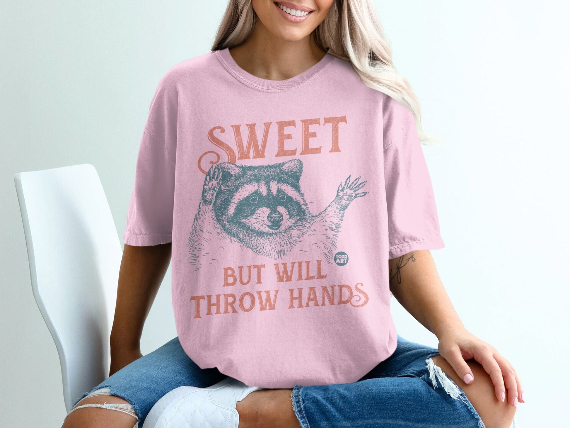 Pink t-shirt with raccoon graphic and text "SWEET BUT WILL THROW HANDS".