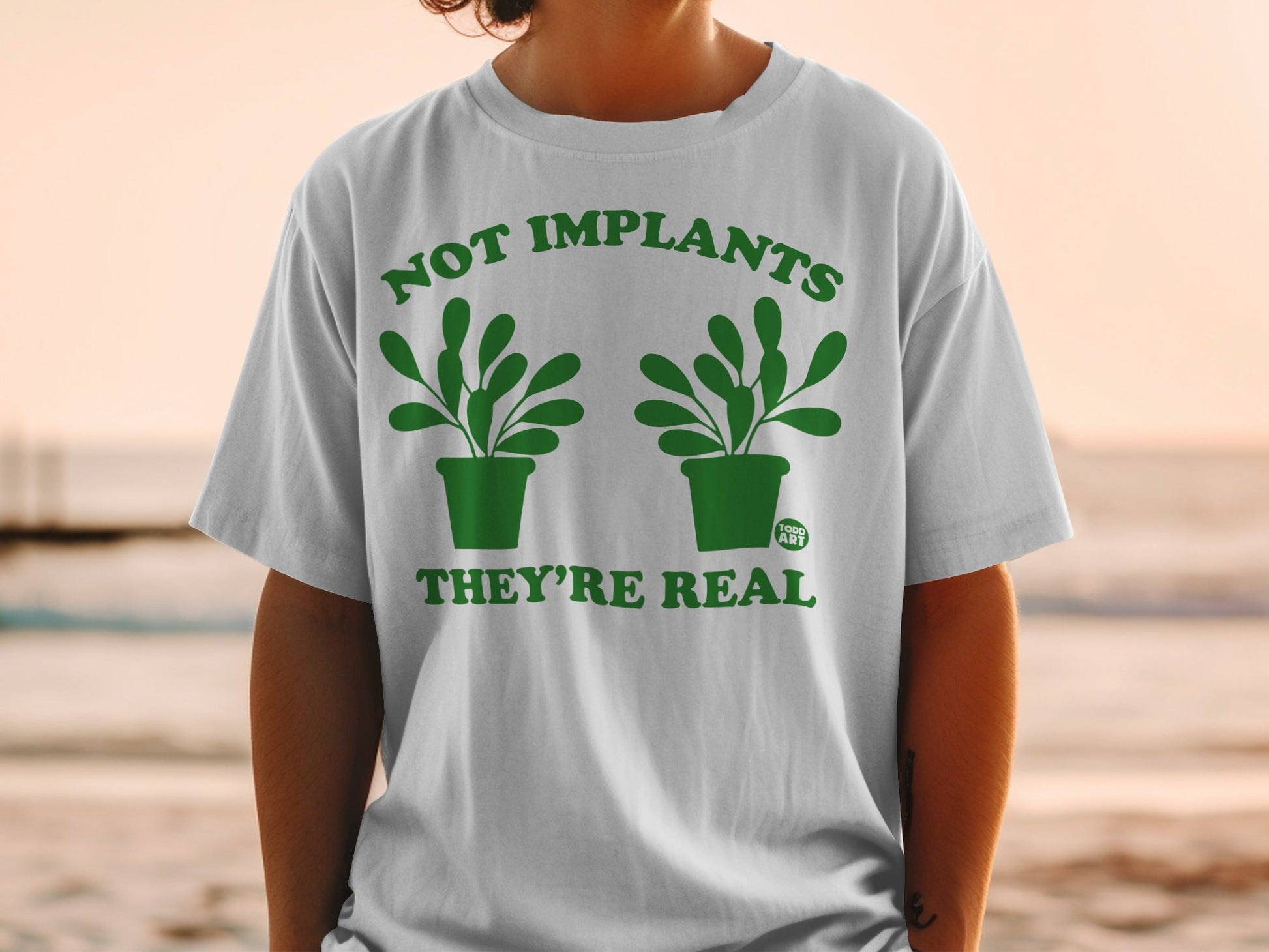 White t-shirt with green text and potted plants reading "NOT IMPLANTS THEY'RE REAL".