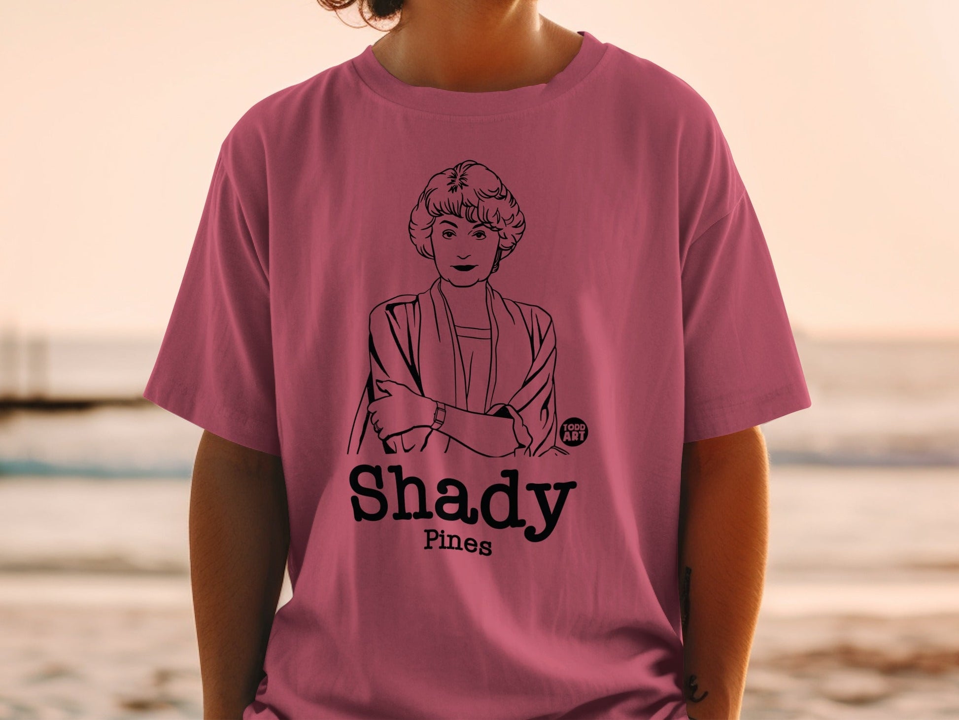 Person wearing a pink t-shirt with a black illustration of a woman and "Shady Pines" text.