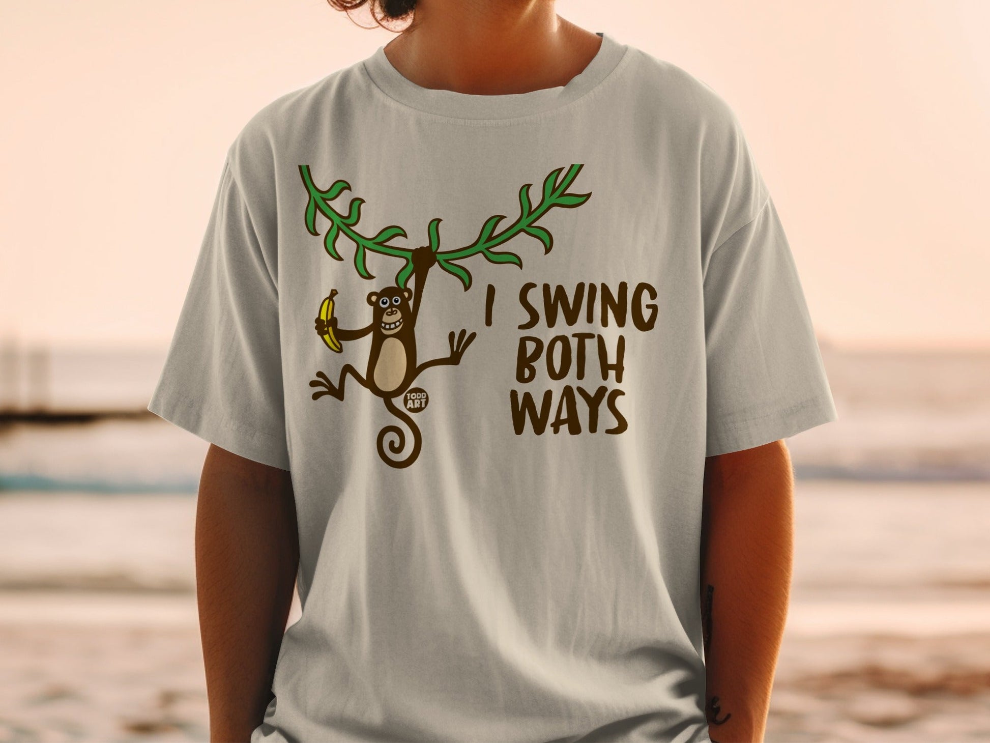 T-shirt with brown monkey swinging from vine holding banana, text "I SWING BOTH WAYS".