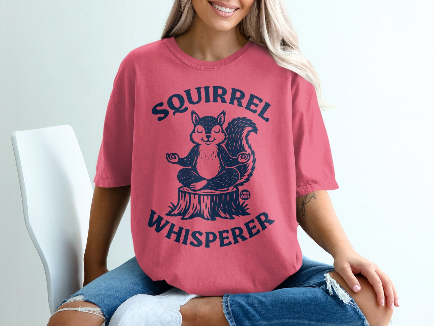 Pink t-shirt with a meditating squirrel on a tree stump, text reads "Squirrel Whisperer".