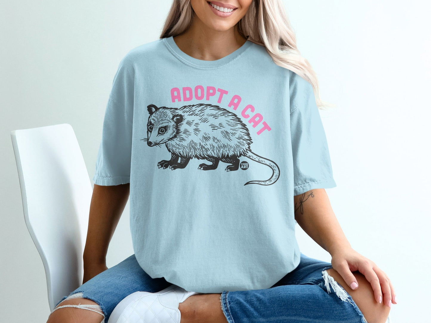 Light blue t-shirt with "Adopt a Cat" text and a stylized opossum illustration.