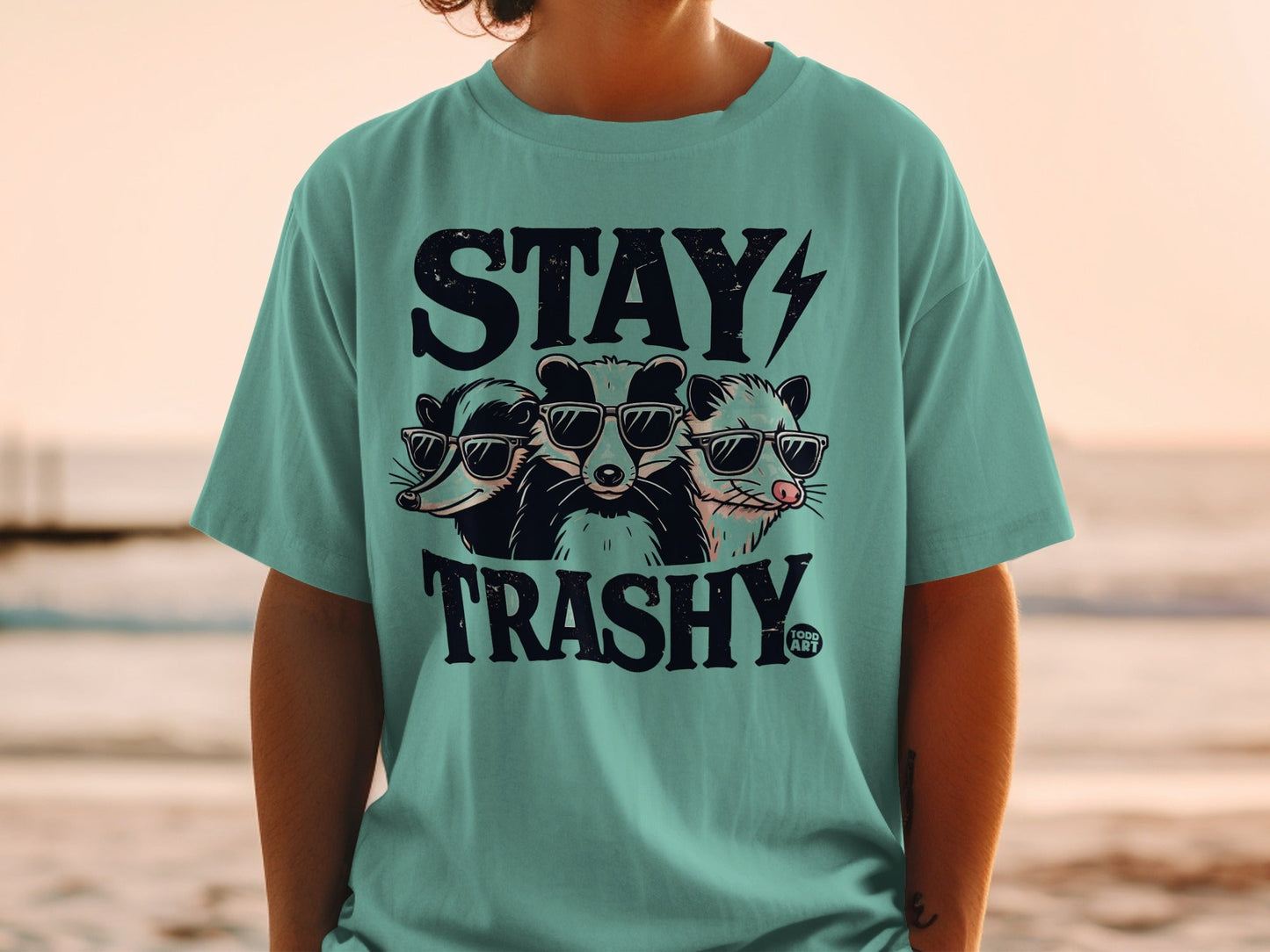 Three raccoons in sunglasses on a teal t-shirt with "STAY TRASHY" text.