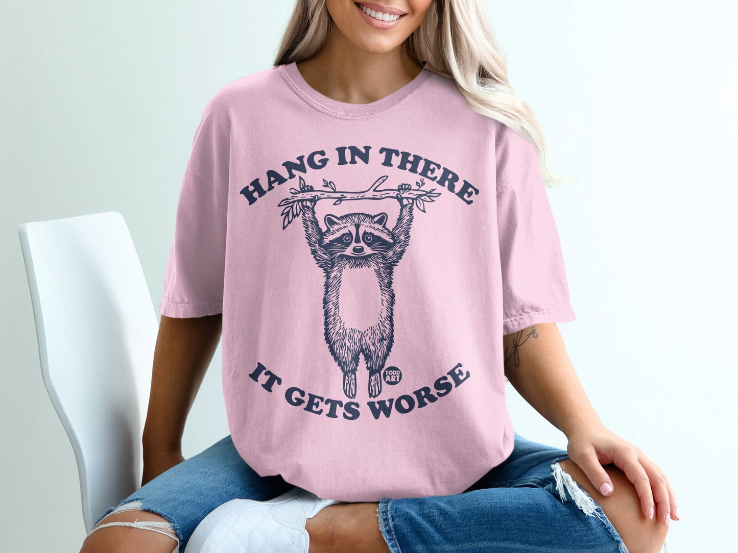 Pink t-shirt with raccoon hanging from branch, text "Hang in there it gets worse".