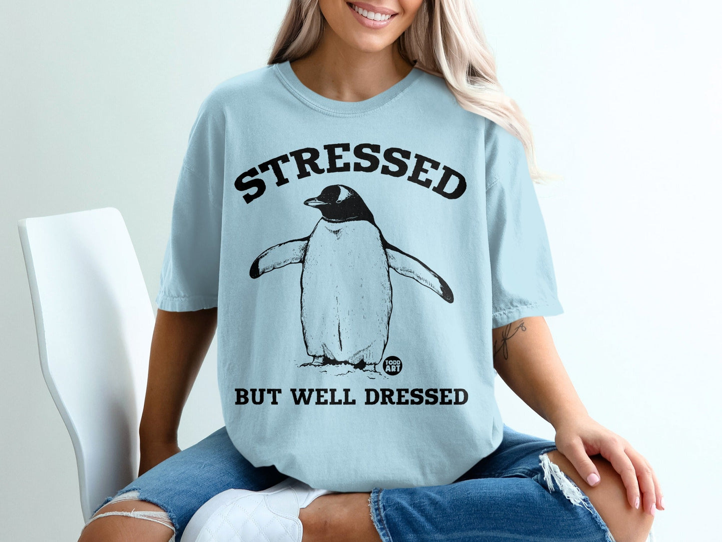 Light blue t-shirt with a penguin graphic and text "STRESSED BUT WELL DRESSED".