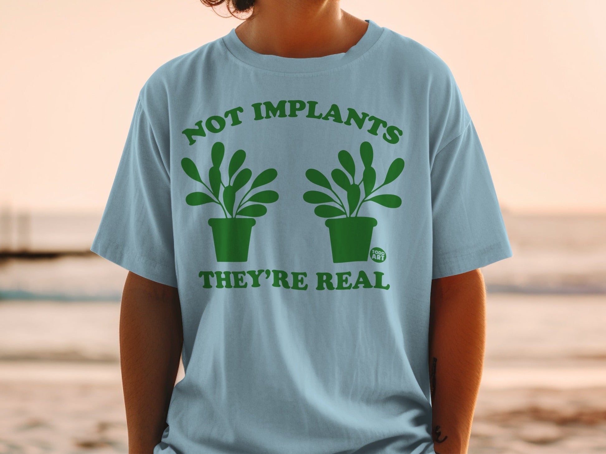 Light blue t-shirt with green graphic reading "Not Implants They're Real" and two potted plants.