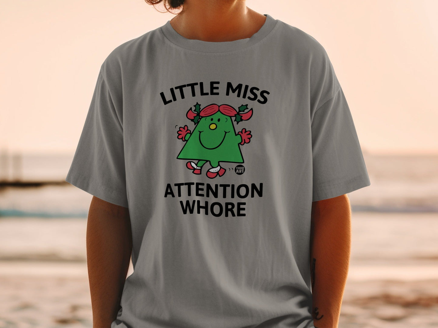 Grey t-shirt with a cartoon Christmas tree character wearing red ribbons and reading "Little Miss Attention Whore."