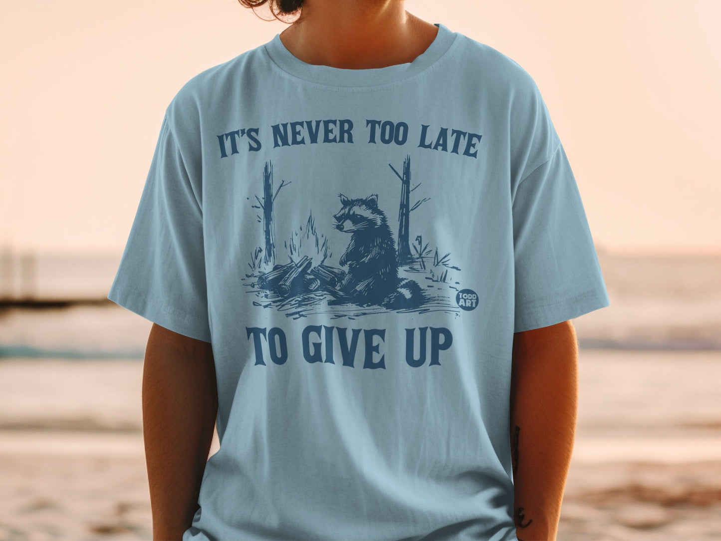 Light blue t-shirt with raccoon and campfire graphic, "It's Never Too Late To Give Up".