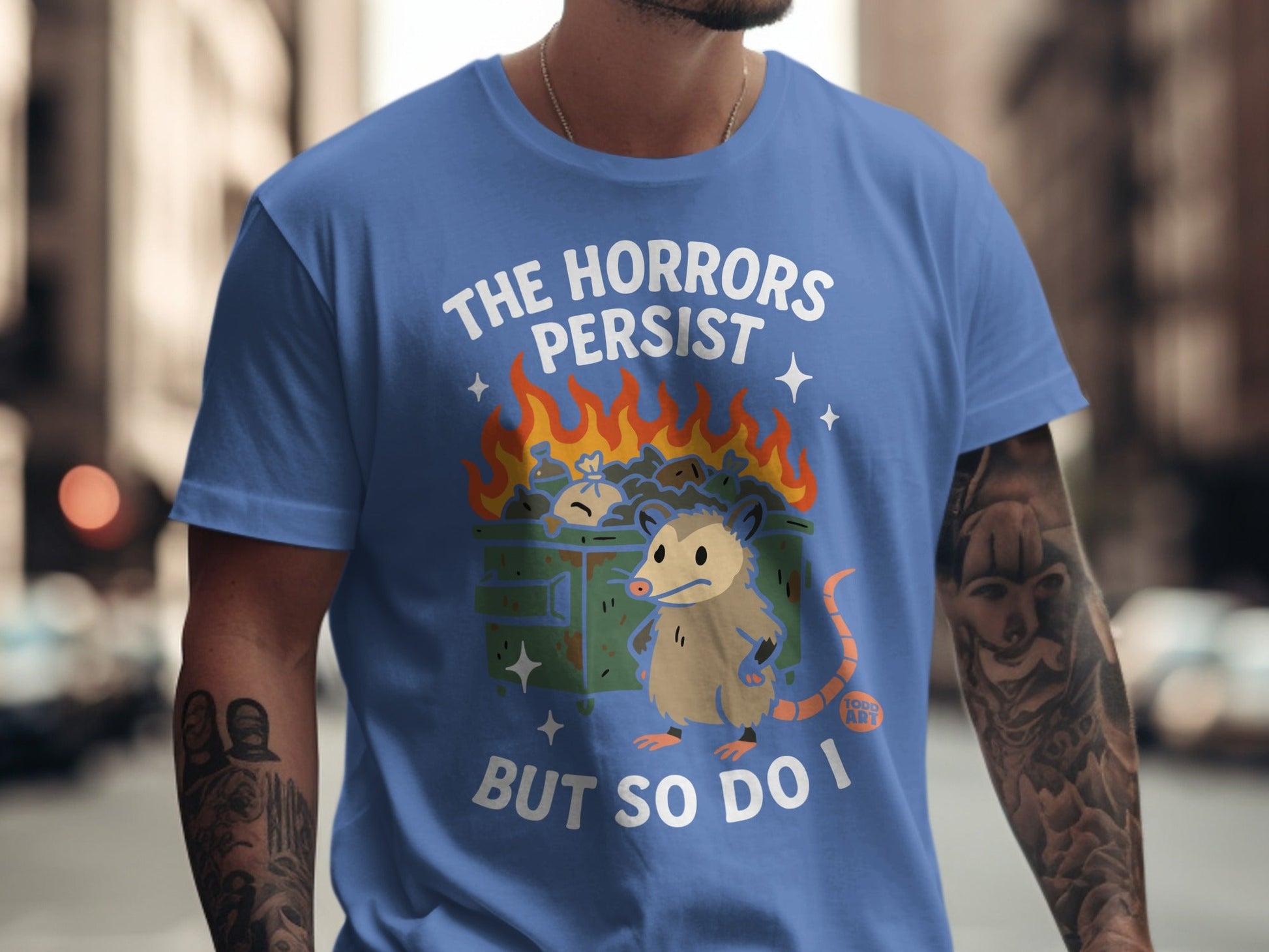 Blue t-shirt with opossum in a burning trash can graphic and text "THE HORRORS PERSIST BUT SO DO I".