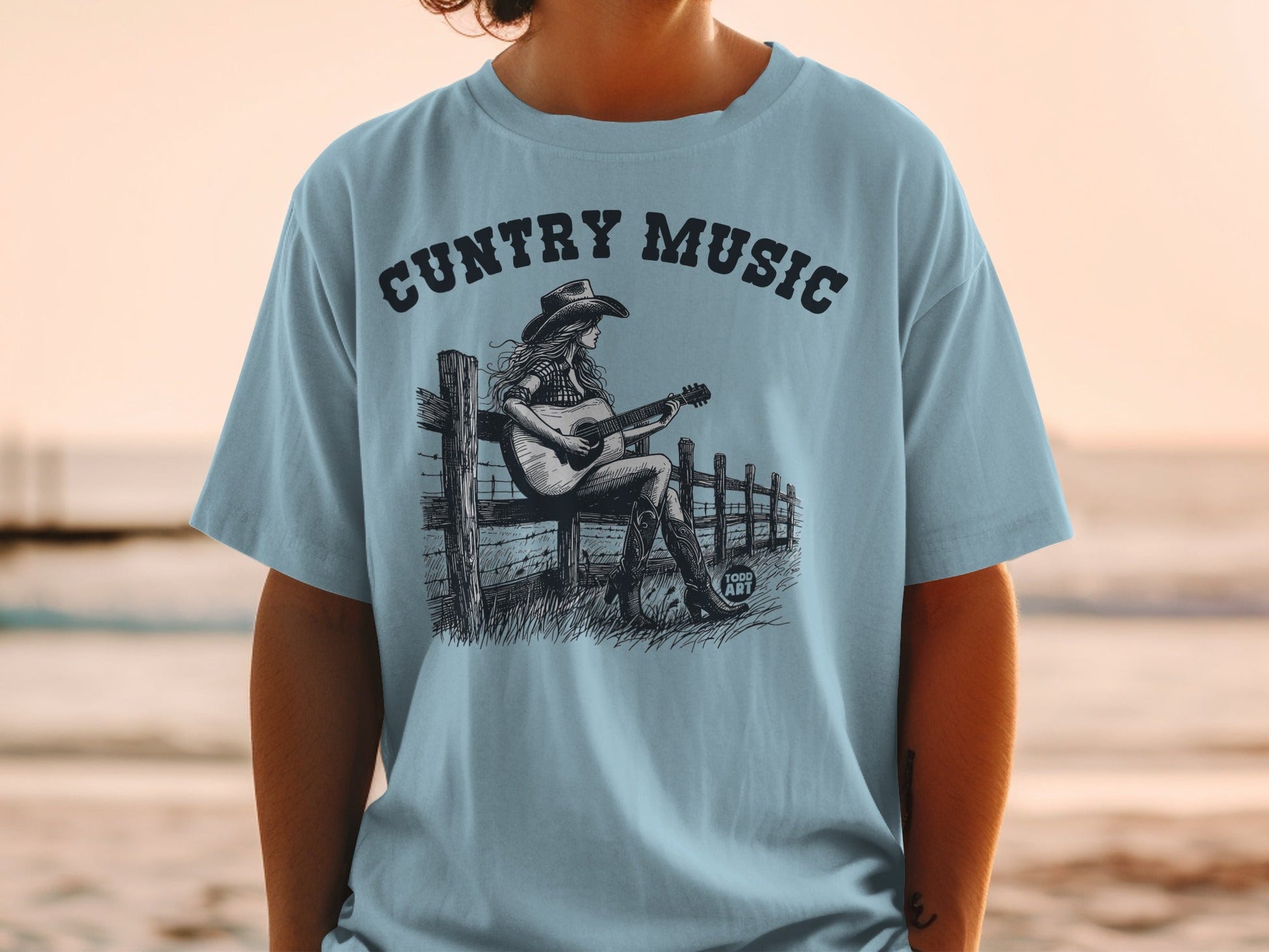 Light blue t-shirt with "Country Music" and woman playing guitar on fence illustration.