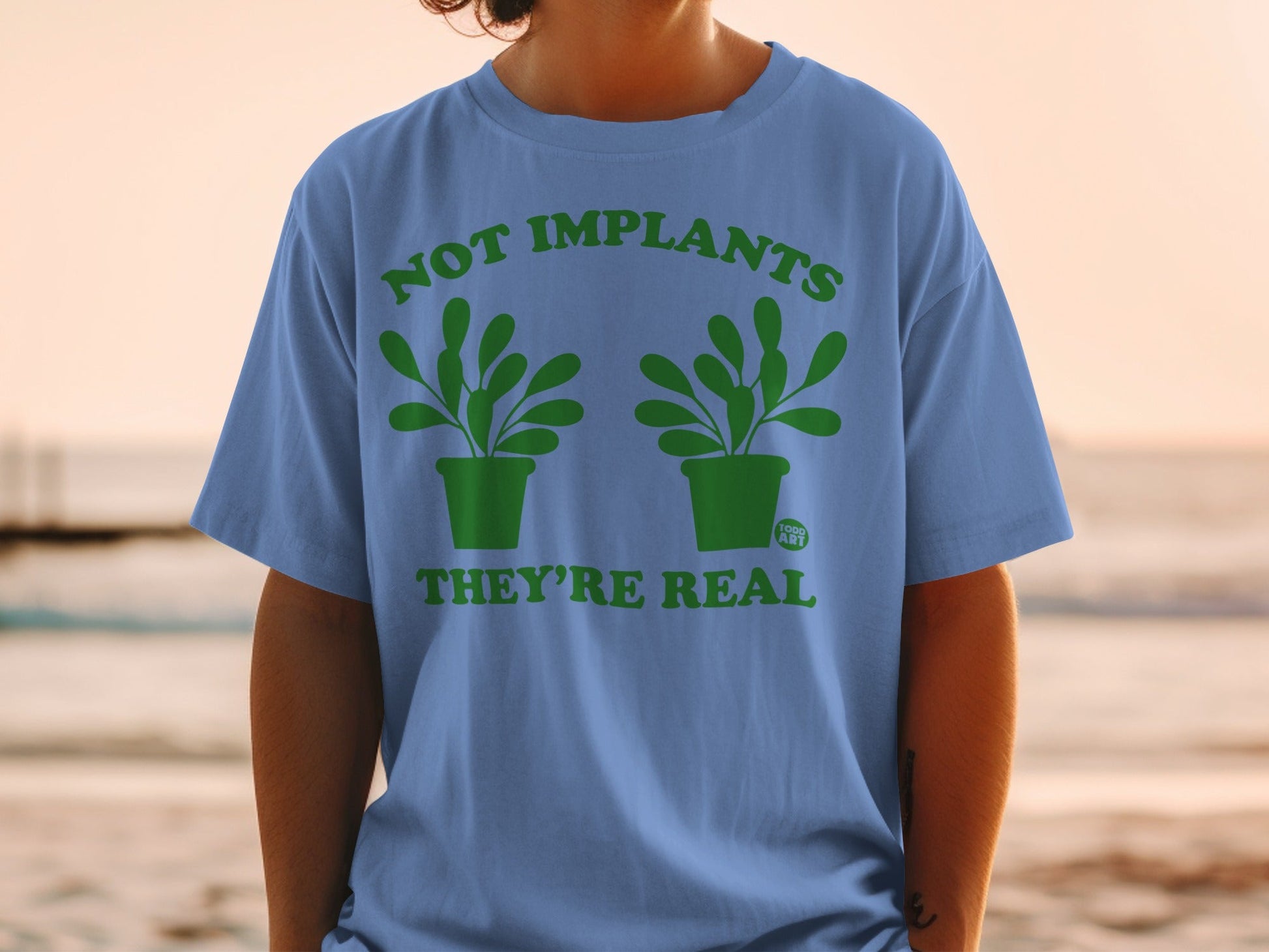 Blue t-shirt with green text and plant graphics reading "NOT IMPLANTS THEY'RE REAL".