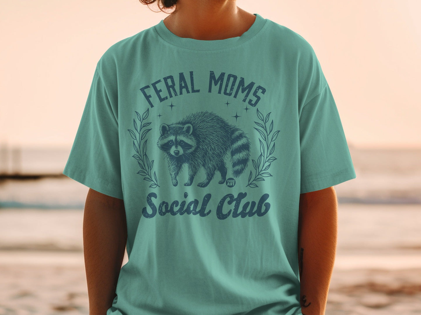 Teal t-shirt with "Feral Moms Social Club" and a raccoon graphic.