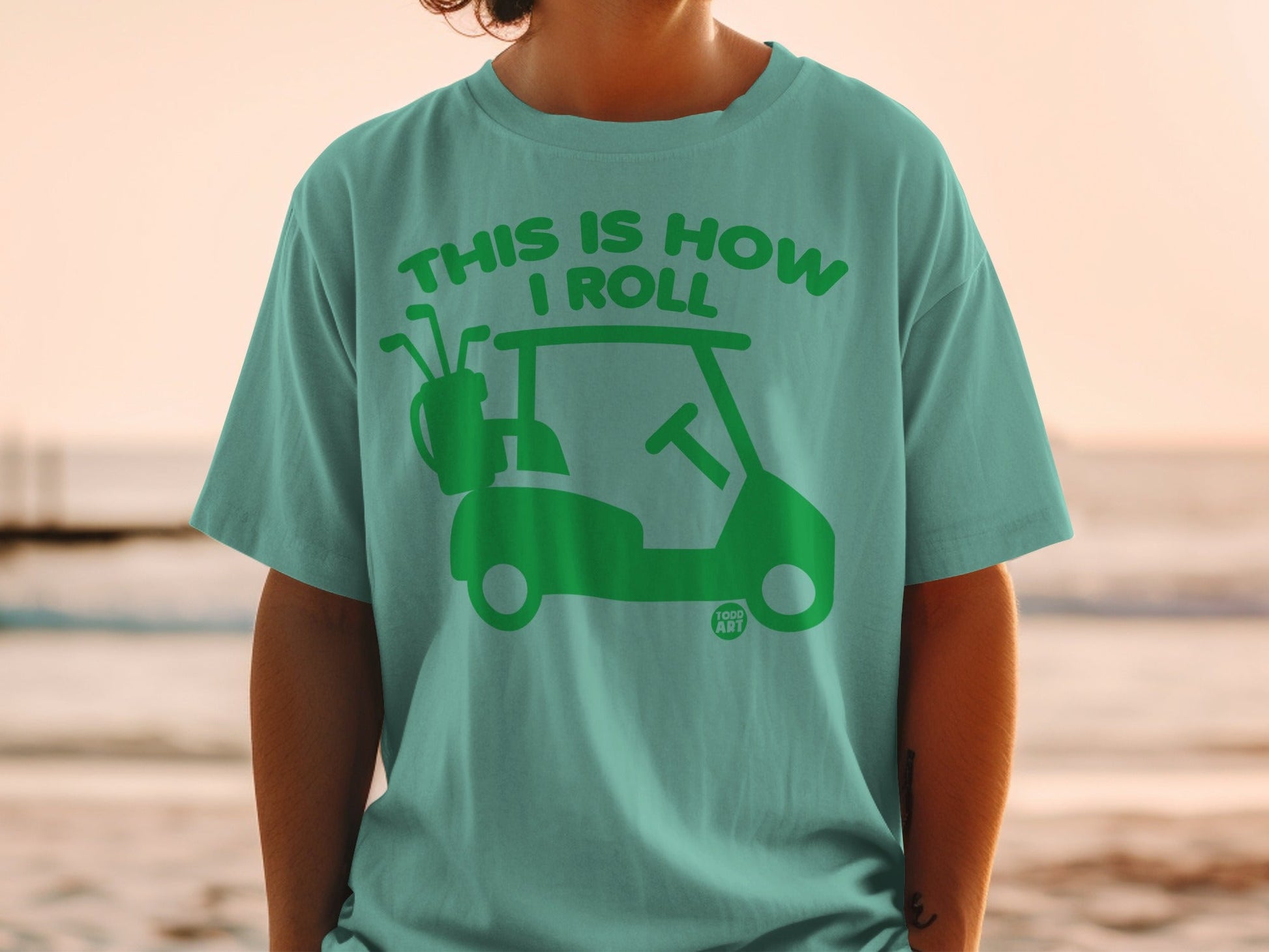 Teal t-shirt with a green golf cart and clubs graphic, text reads "This is how I roll".