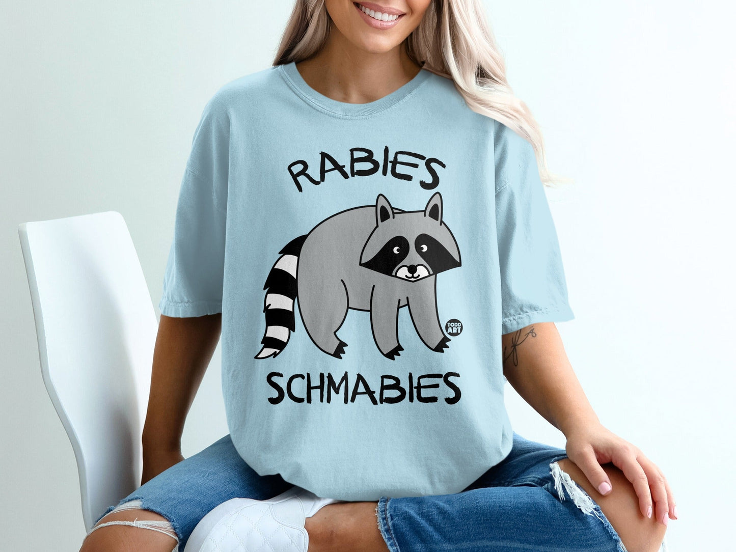 Light blue t-shirt with raccoon graphic and text "RABIES SCHMABIES".