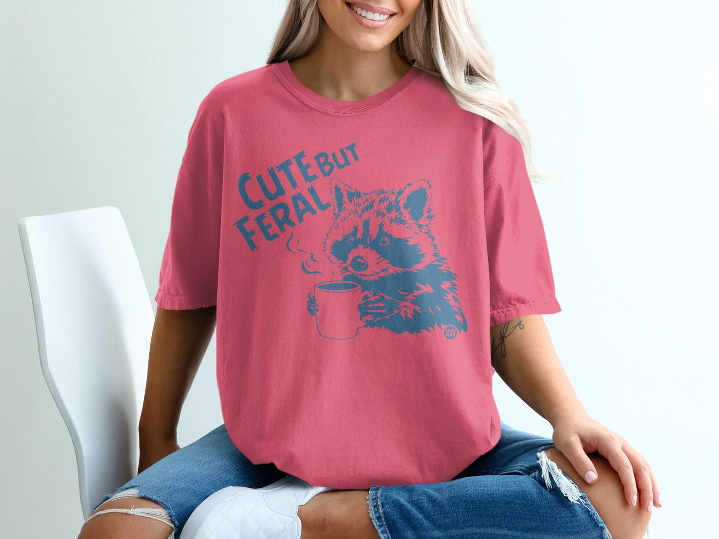 Pink t-shirt with raccoon drinking coffee graphic and text "Cute But Feral".