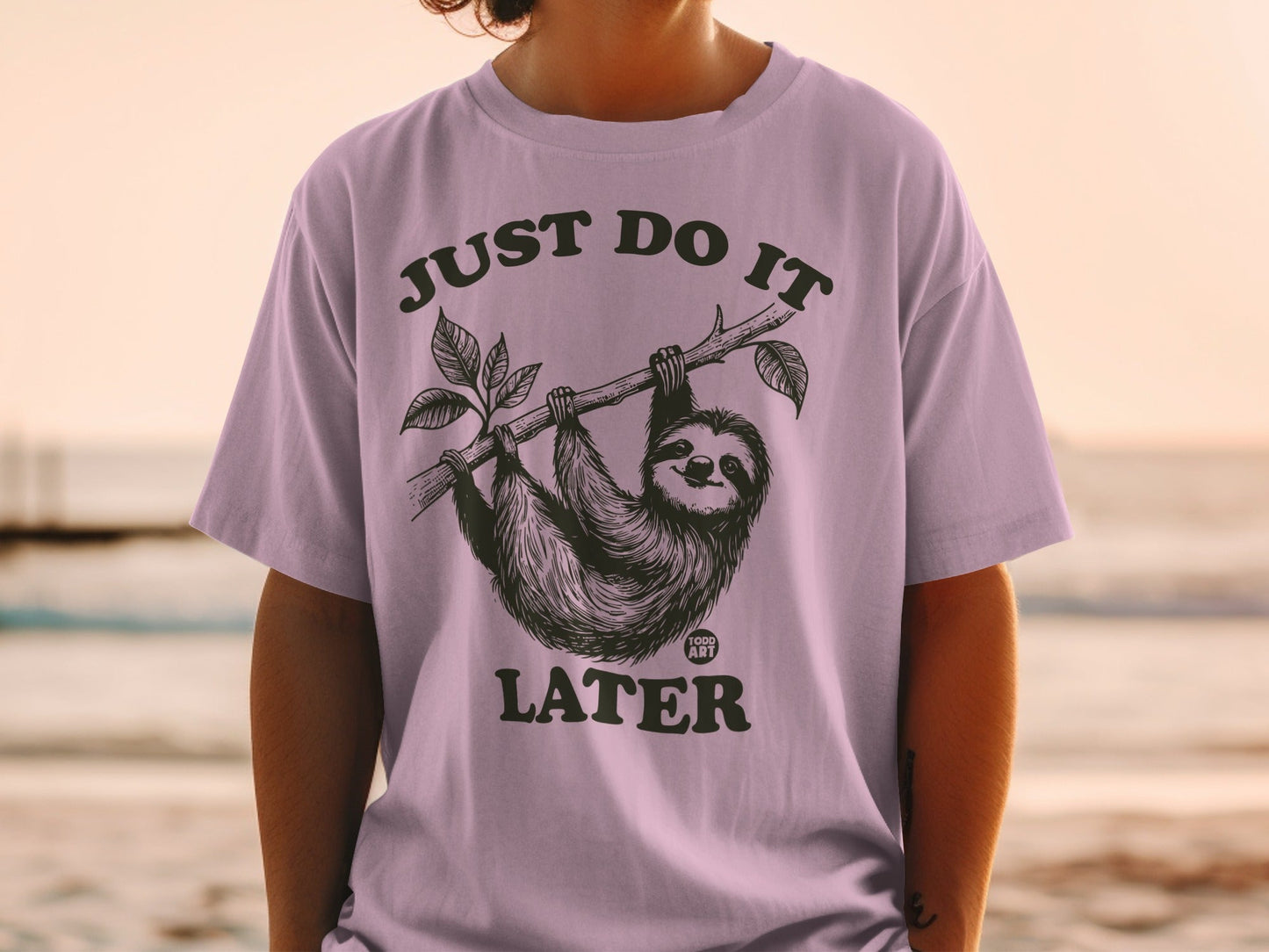 Sloth hanging from branch on purple t-shirt, text reads "JUST DO IT LATER".