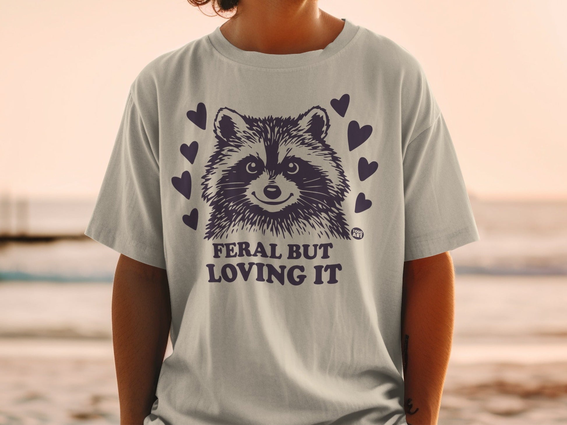 Grey t-shirt with a smiling raccoon graphic and "FERAL BUT LOVING IT" text.
