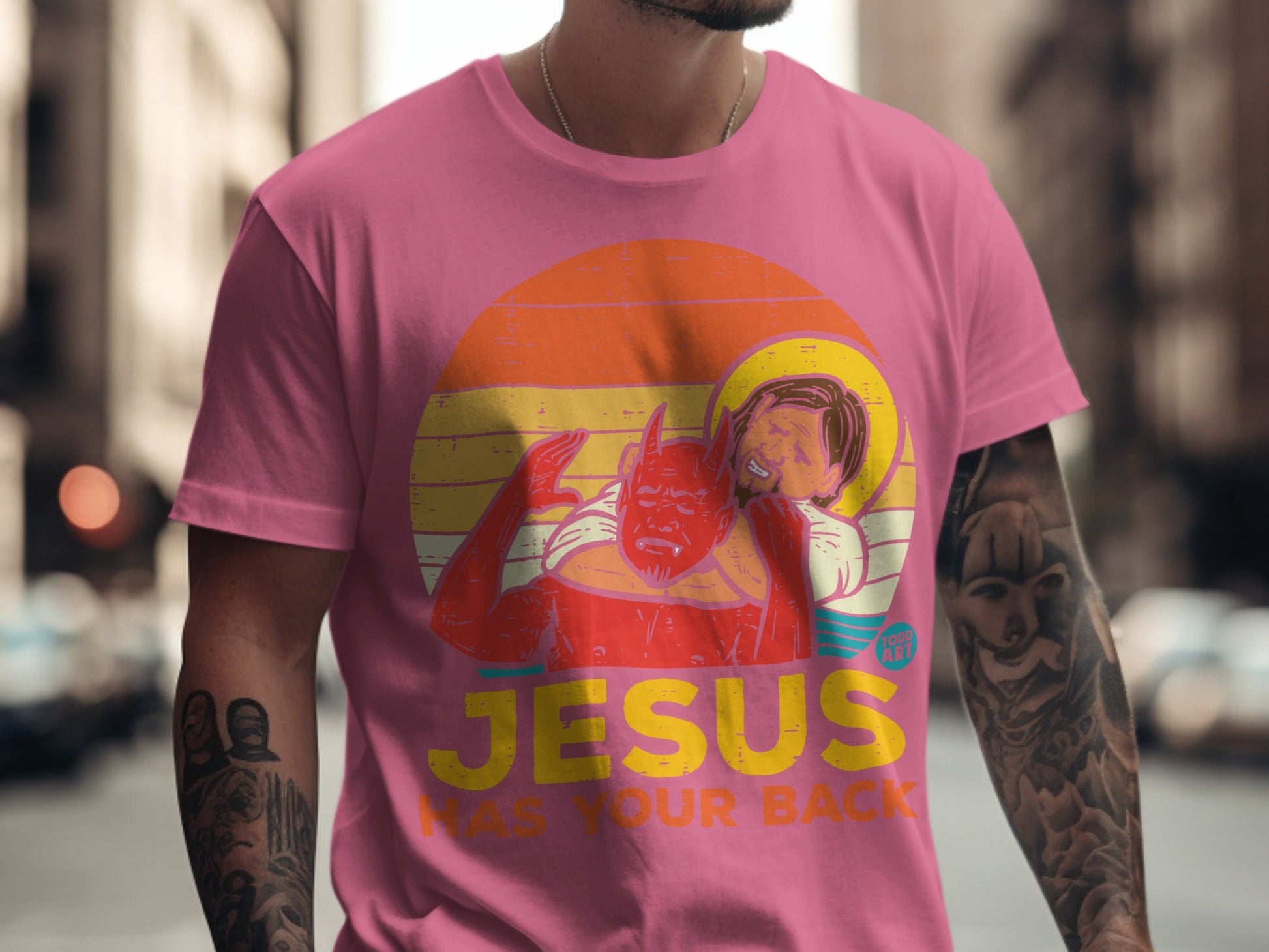 Pink t-shirt with graphic of Jesus and devil, "Jesus Has Your Back" text.