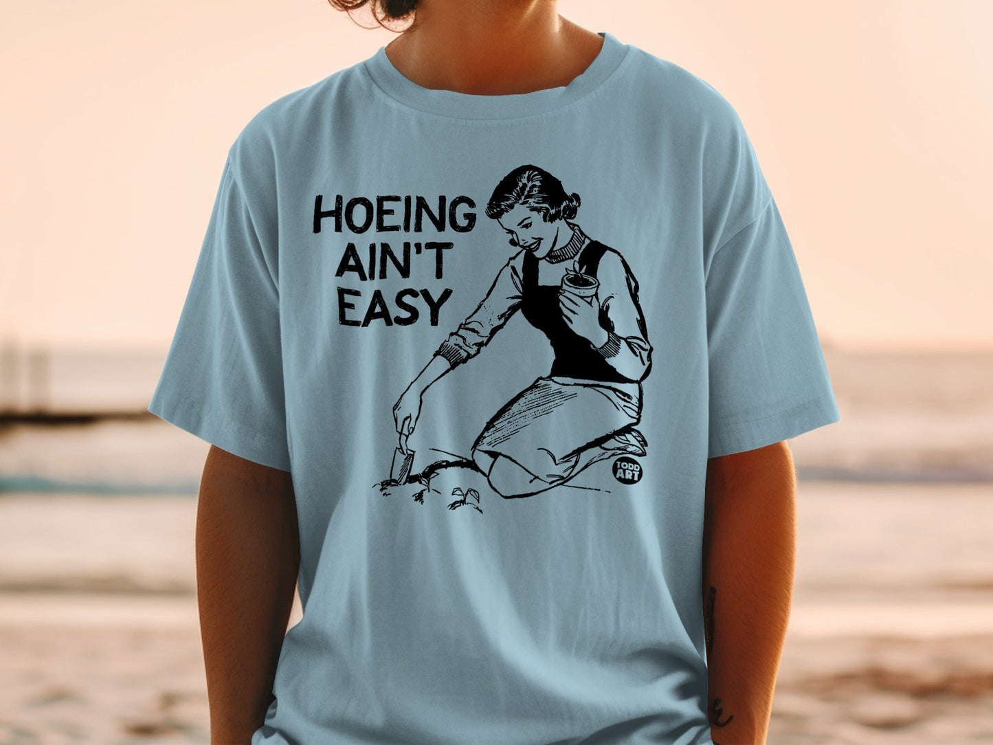 Black text "Hoing Ain't Easy" above woman tending garden, wearing light blue t-shirt.