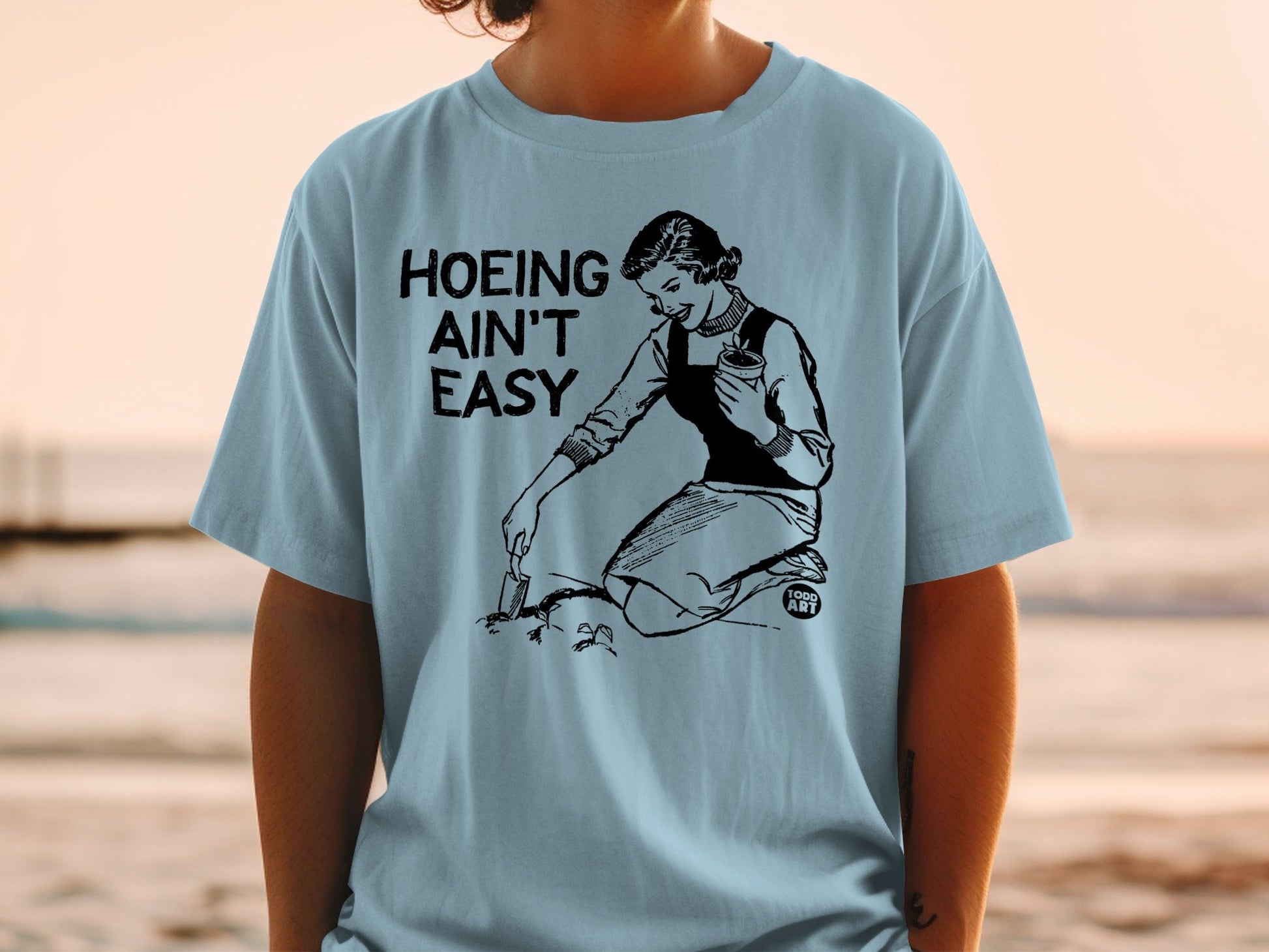 Black text "Hoing Ain't Easy" above woman tending garden, wearing light blue t-shirt.