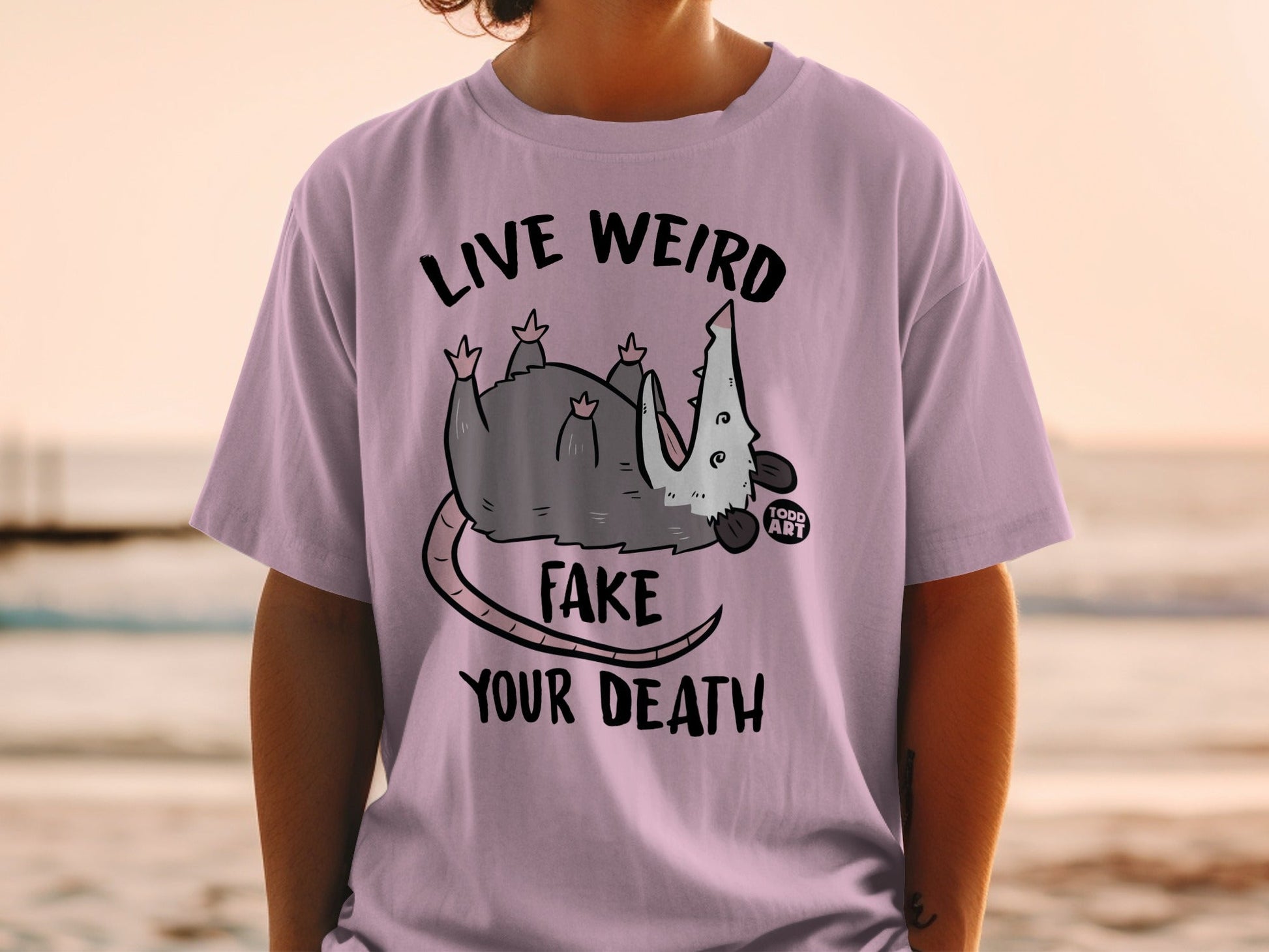 Purple t-shirt with a possum graphic and text "LIVE WEIRD FAKE YOUR DEATH".