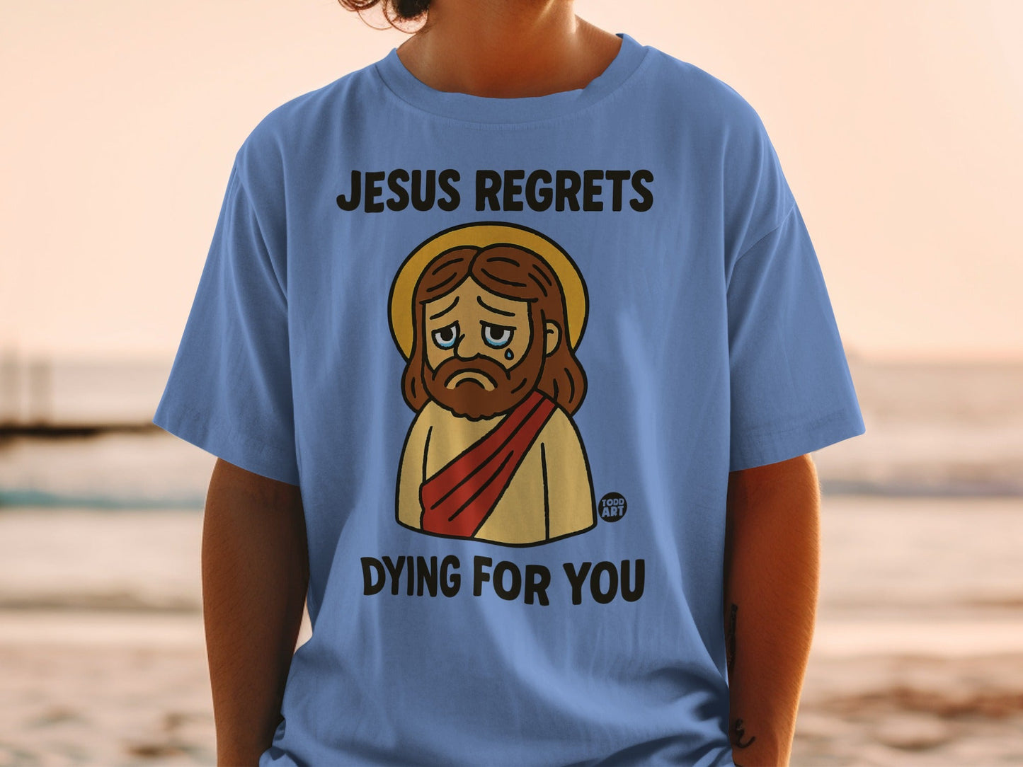 Blue t-shirt with cartoon Jesus wearing halo, crying, text "JESUS REGRETS DYING FOR YOU".