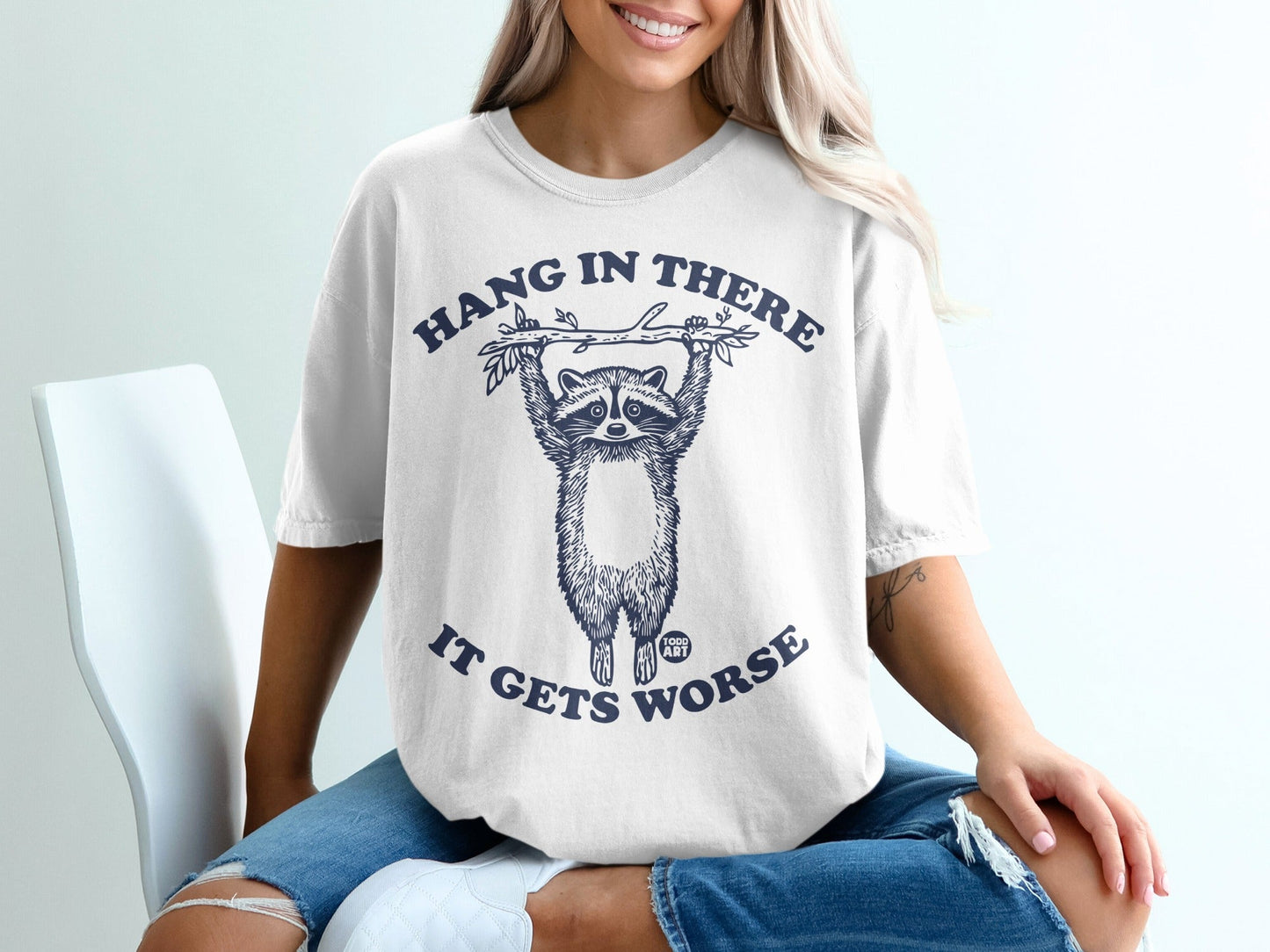 White t-shirt with raccoon hanging from a branch, text: "Hang in there, it gets worse."