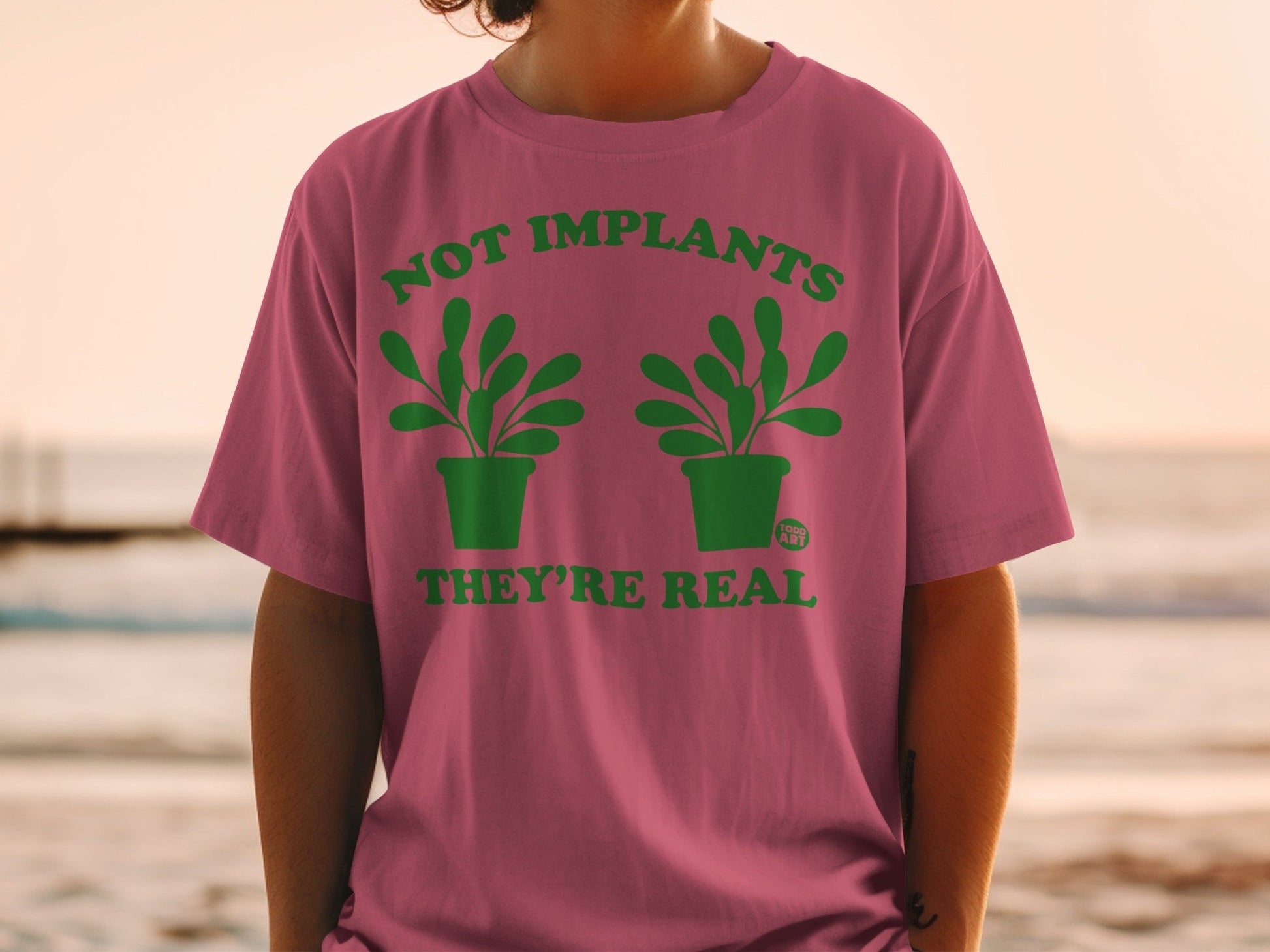 Pink t-shirt with green text and plant graphics: "NOT IMPLANTS THEY'RE REAL".