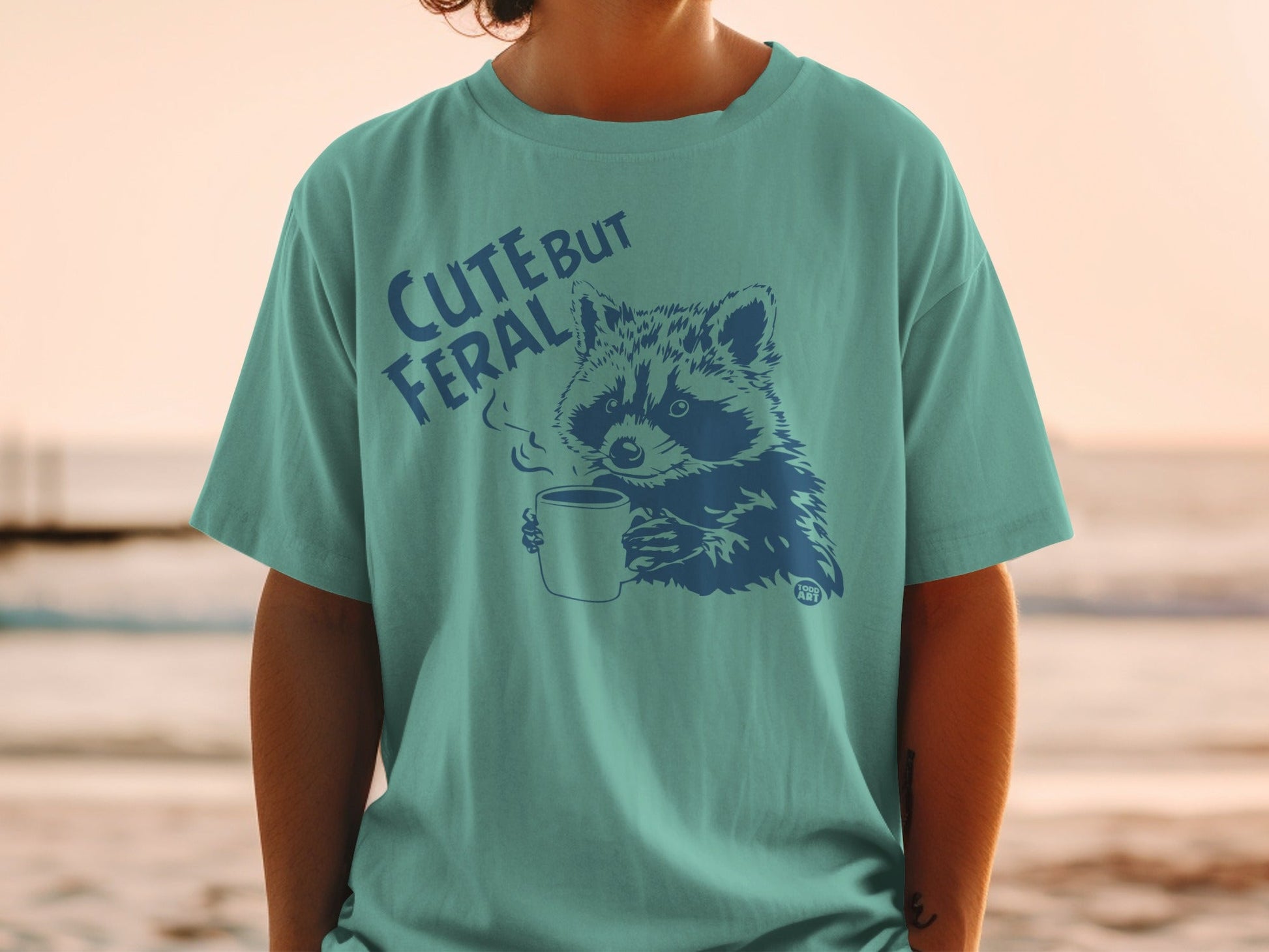 Teal t-shirt with blue raccoon graphic and text "Cute but Feral".