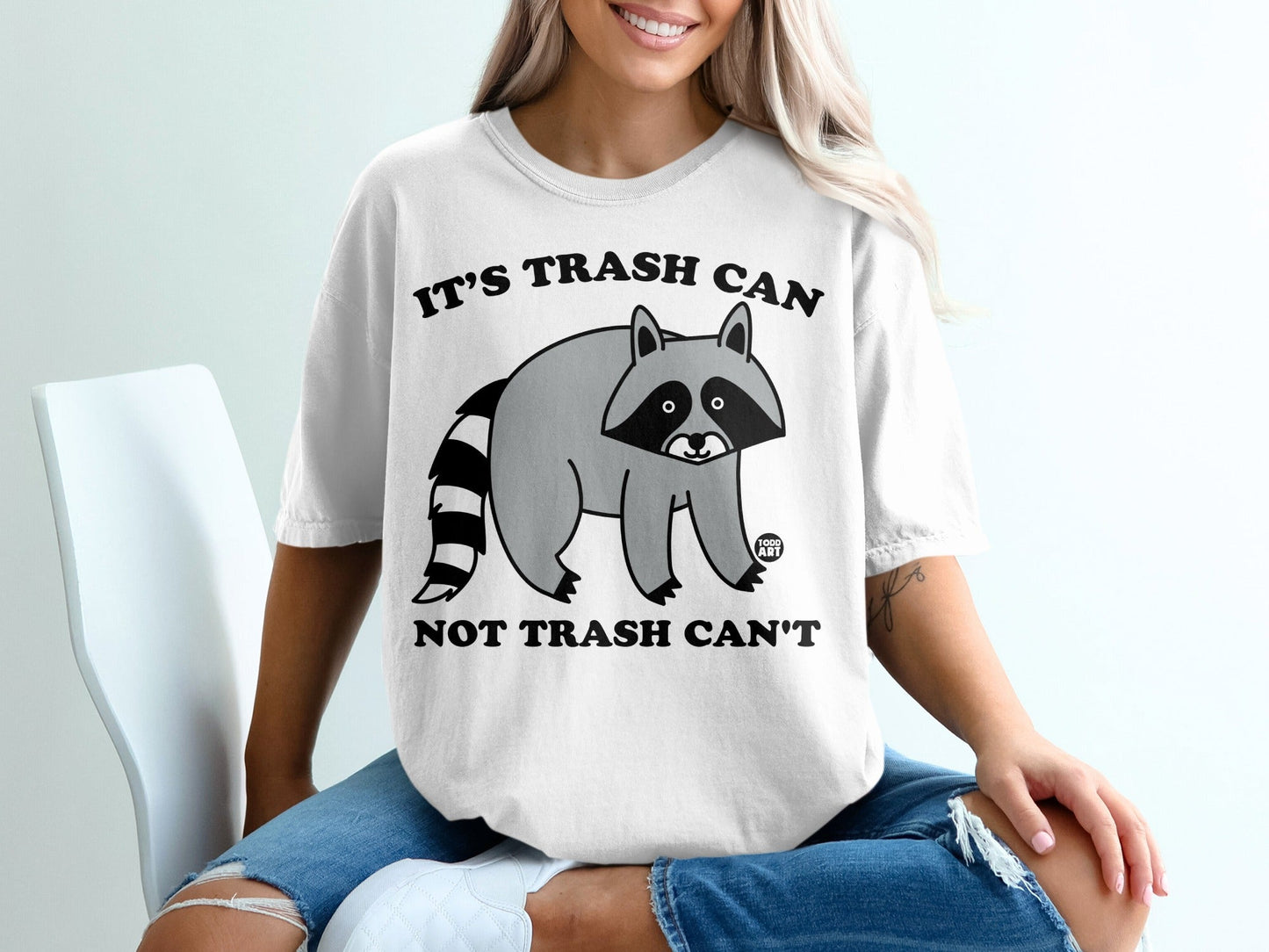 White t-shirt with raccoon graphic and "IT'S TRASH CAN NOT TRASH CAN'T" text.