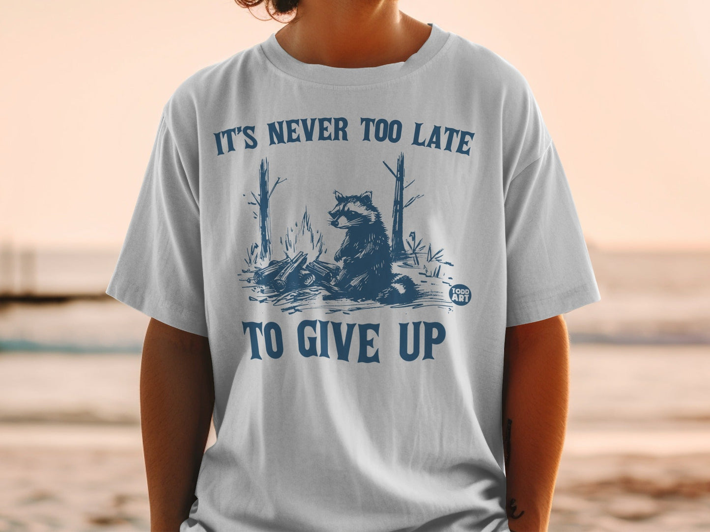 T-shirt graphic with raccoon by campfire, text "IT'S NEVER TOO LATE TO GIVE UP".