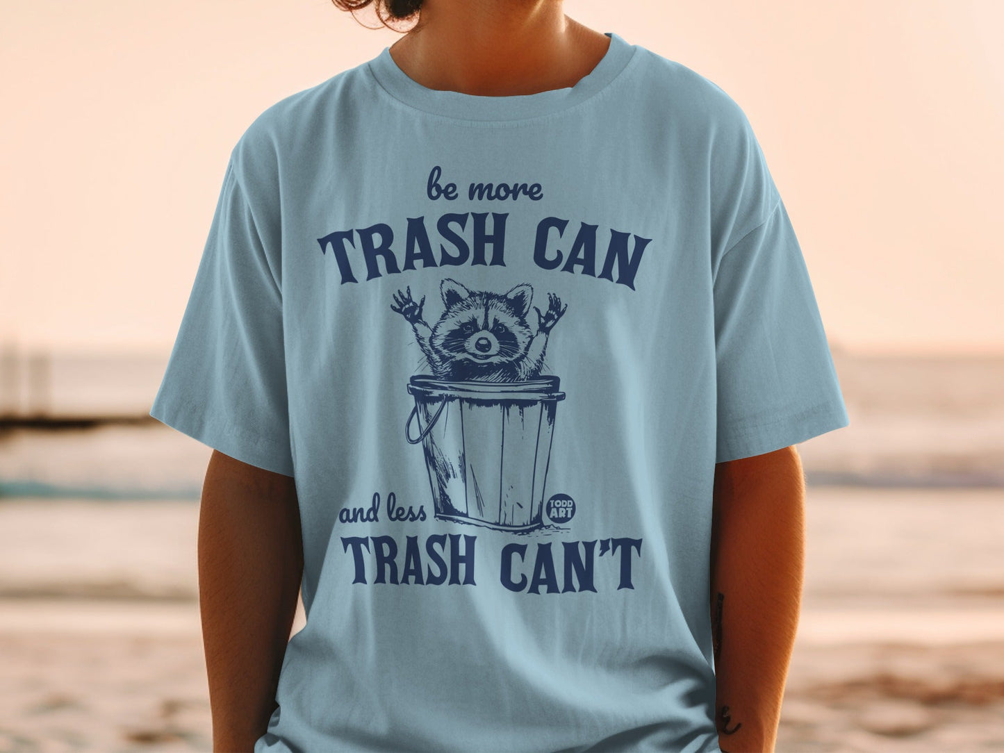 Blue t-shirt with raccoon in trash can graphic and "be more trash can and less trash can't" text.