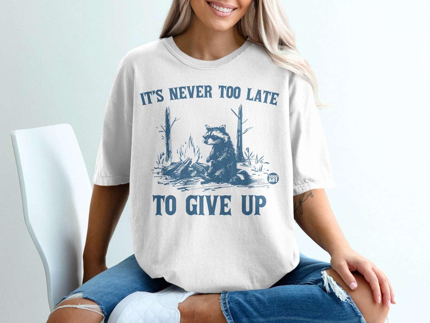 White t-shirt with raccoon and campfire graphic, "It's never too late to give up."