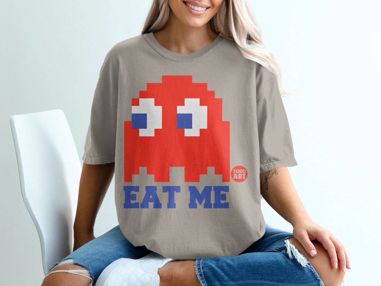 Red pixelated ghost with blue eyes and "EAT ME" text on a tan t-shirt.