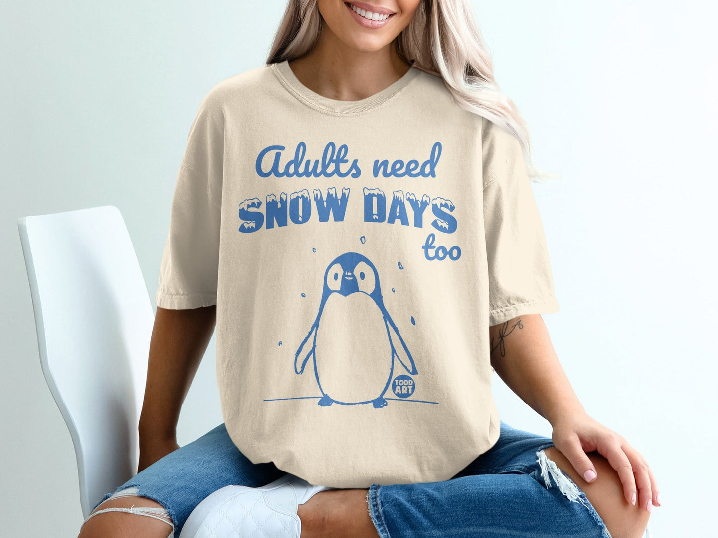 Cream t-shirt with a cartoon penguin and text "Adults need snow days too".