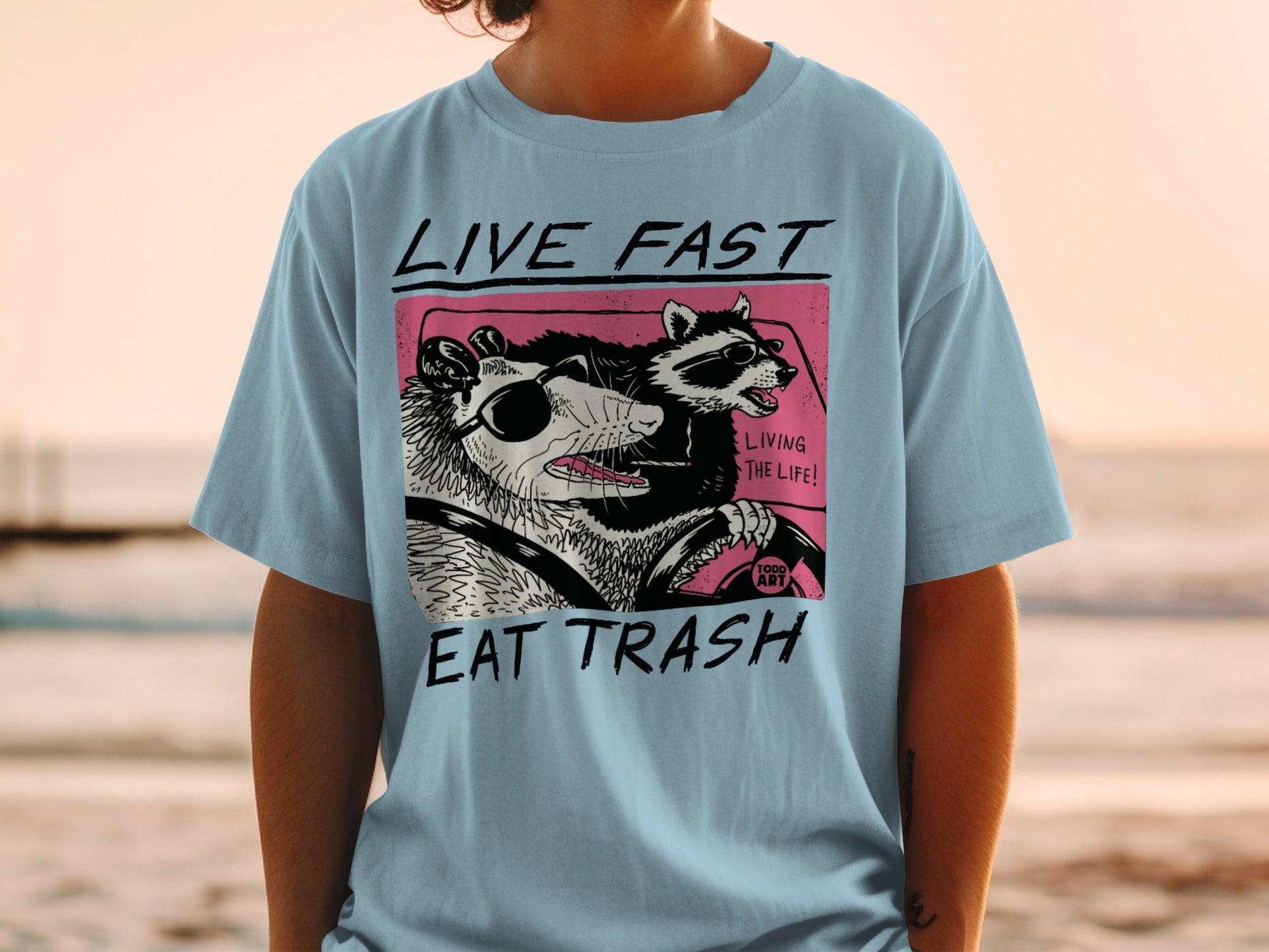 Light blue t-shirt with graphic of raccoon and possum driving, "LIVE FAST EAT TRASH" text.