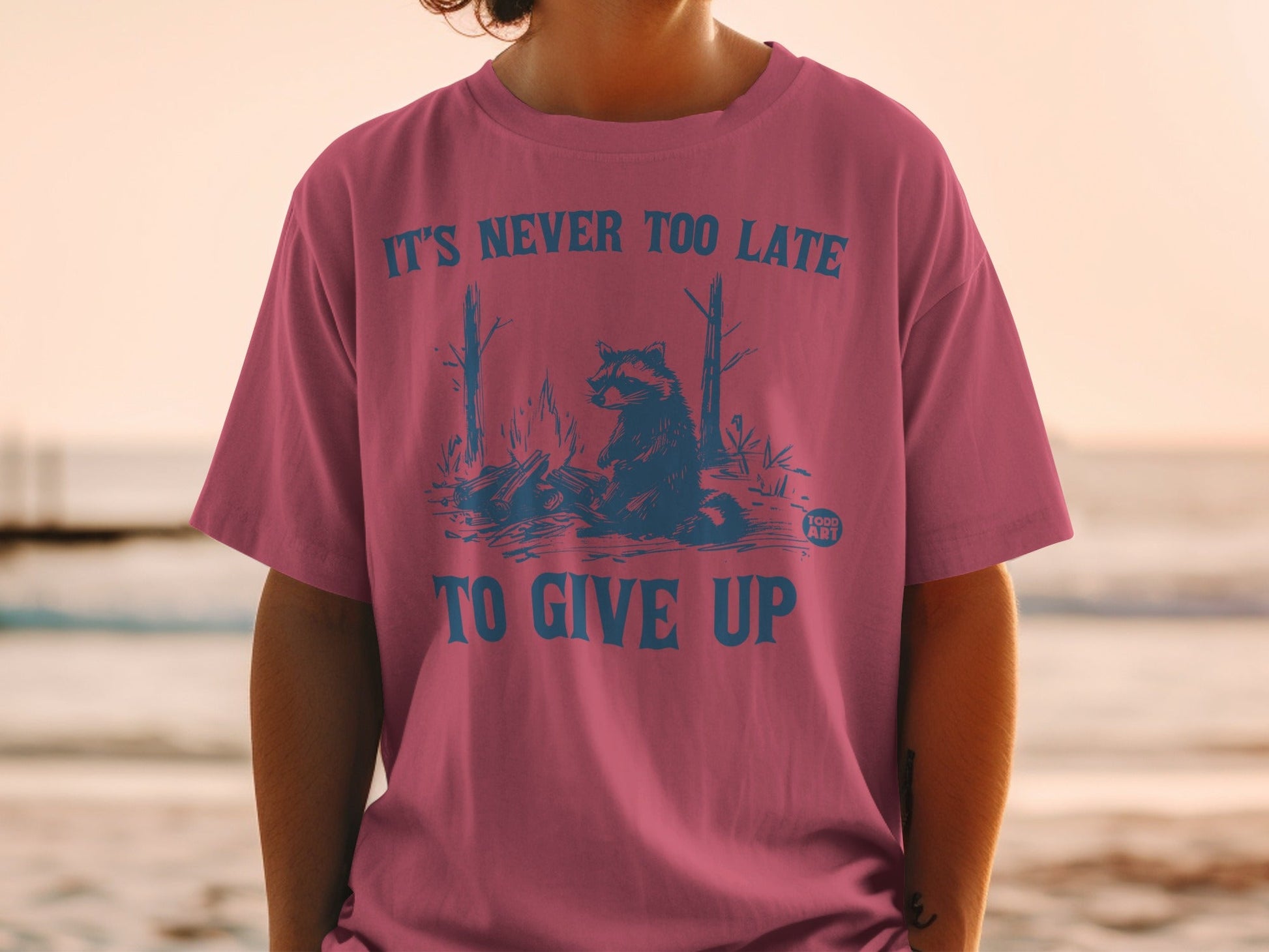 Pink t-shirt with raccoon and campfire graphic and "It's Never Too Late To Give Up" text.