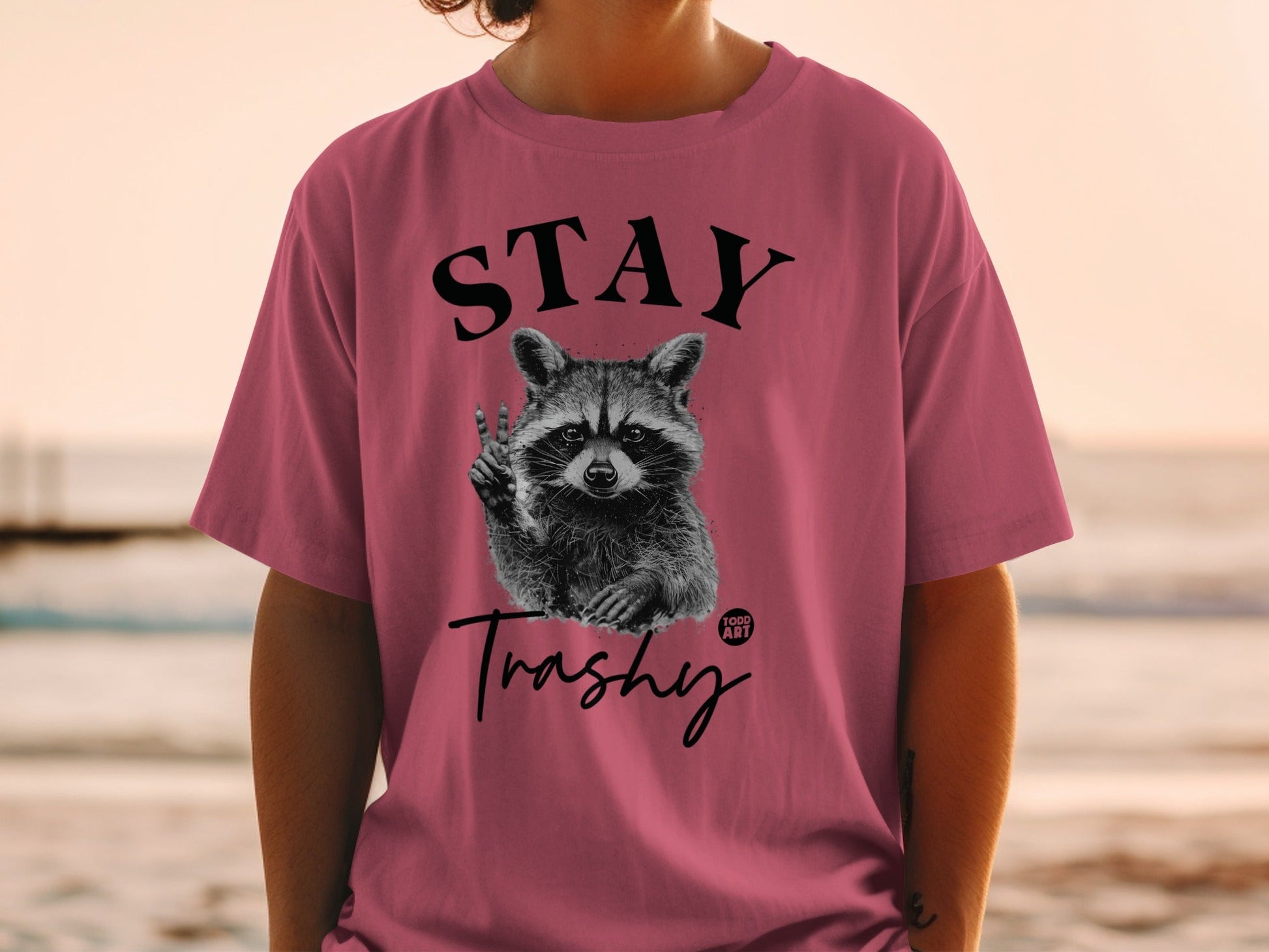 Pink t-shirt with raccoon giving peace sign and "STAY Trashy" text.