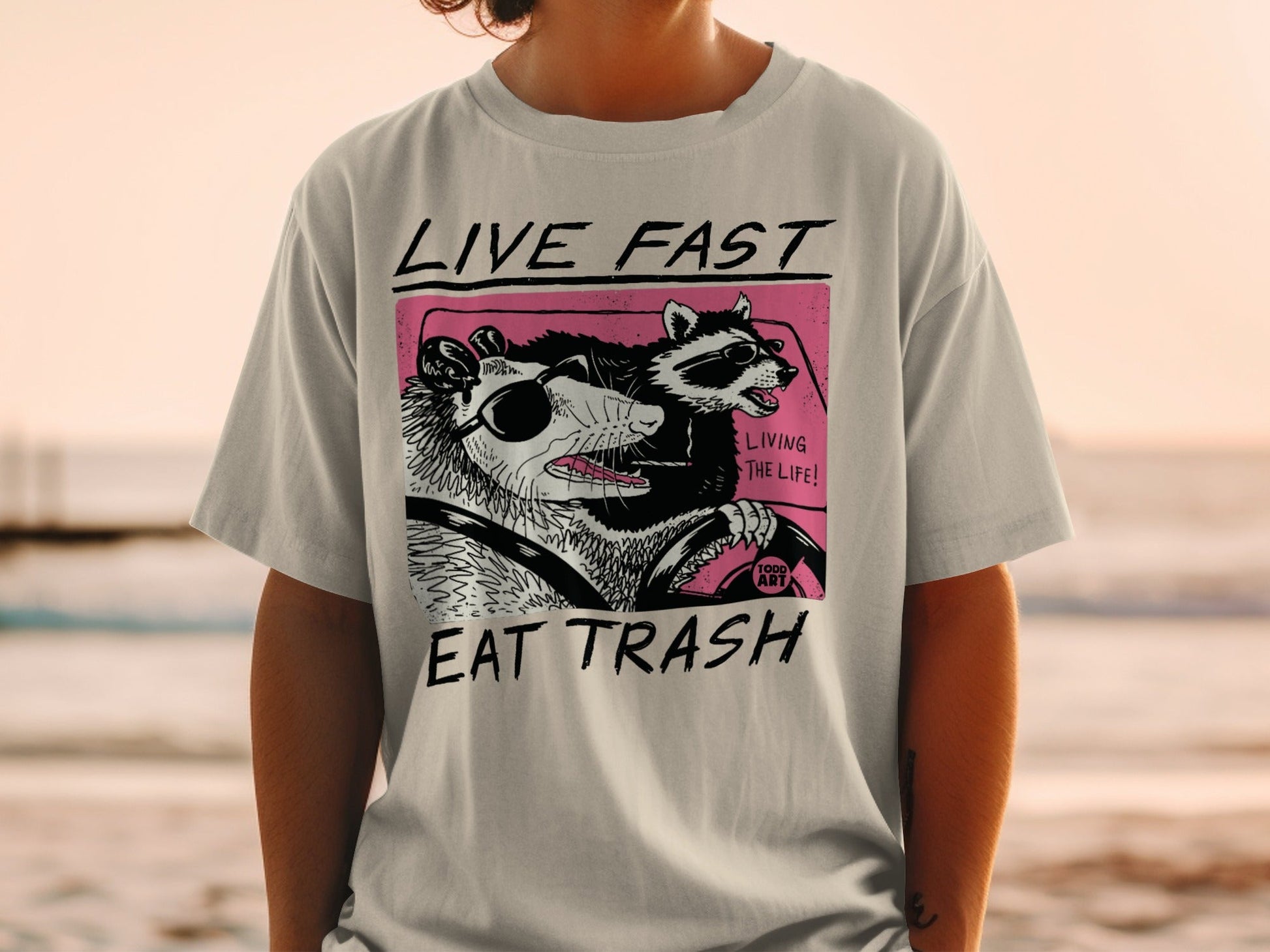 Opossum and raccoon in car, "Live Fast Eat Trash" text on t-shirt.
