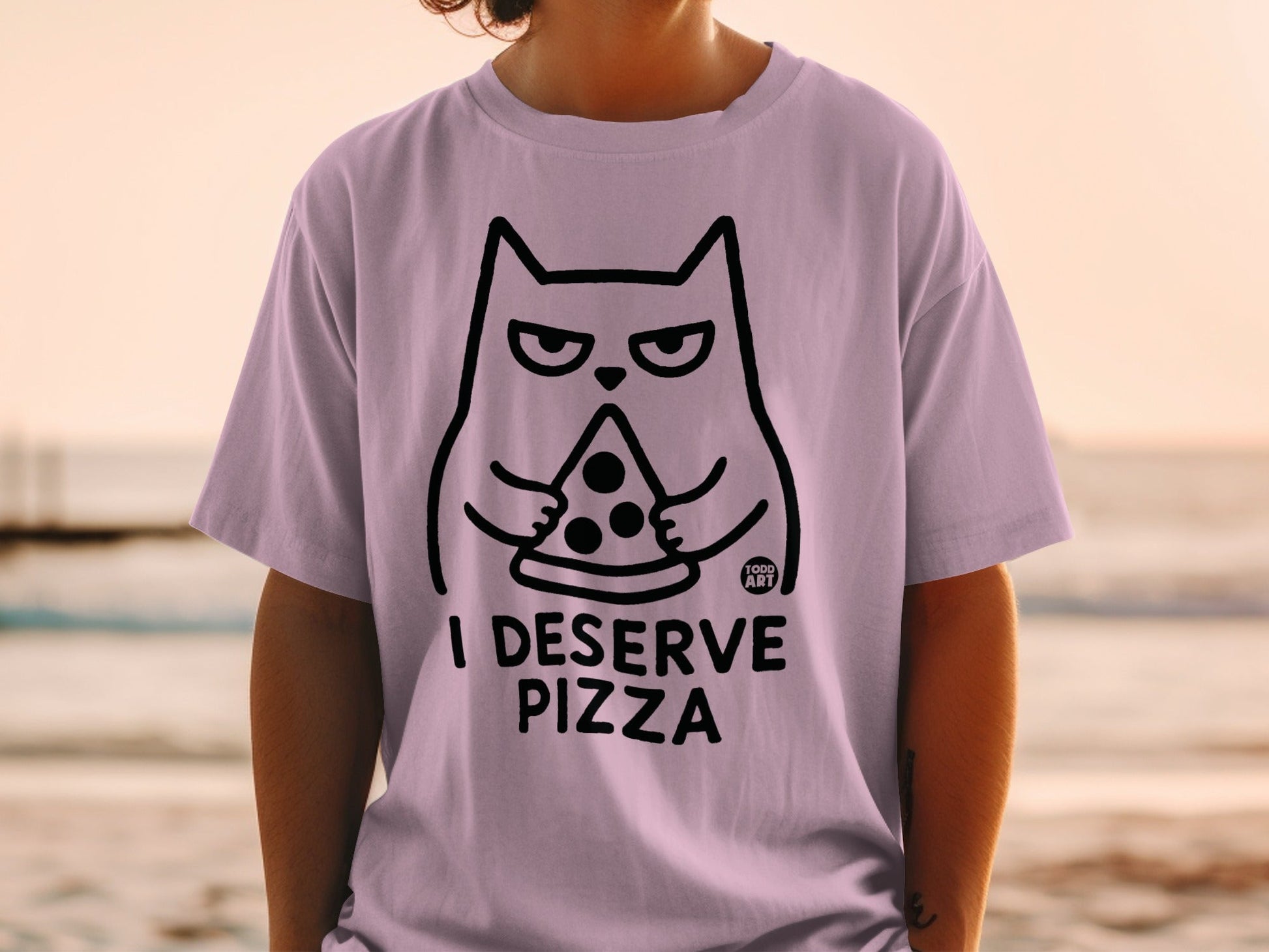 Light purple t-shirt with a black line drawing of a cat holding a slice of pizza and the text "I DESERVE PIZZA".